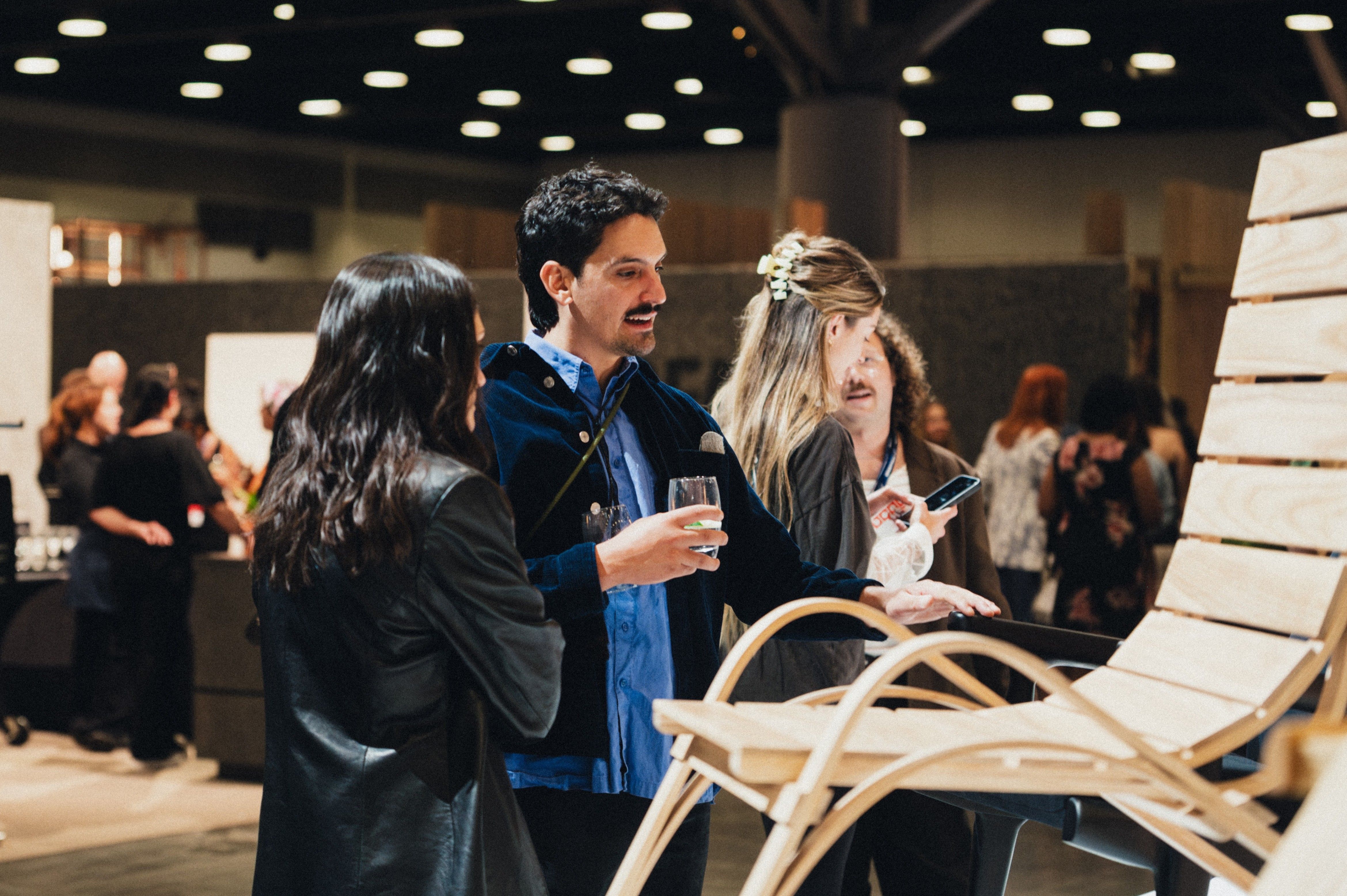 A group of people at an indoor event examining a wooden chair, one person holding a glass and another holding a phone.