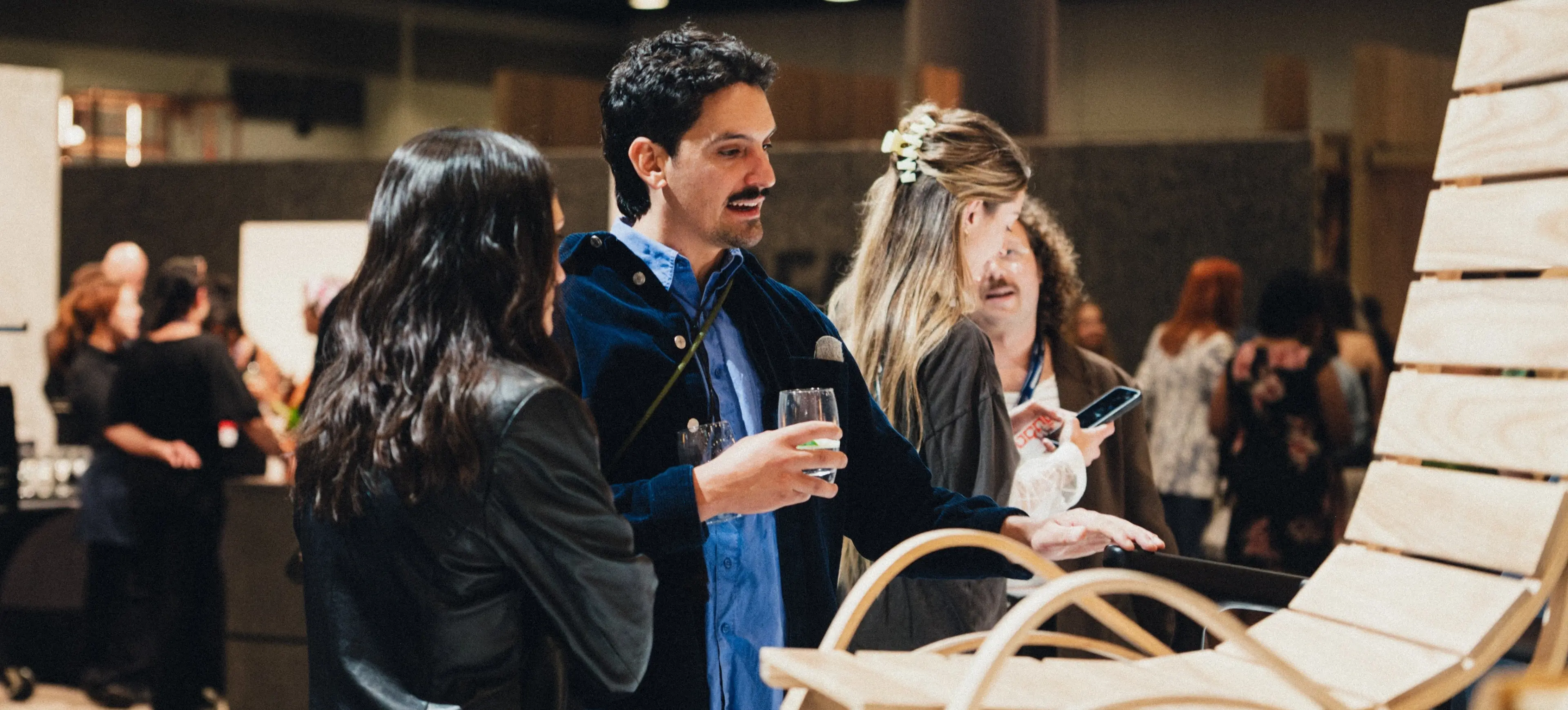 A group of people at an indoor event examining a wooden chair, one person holding a glass and another holding a phone.