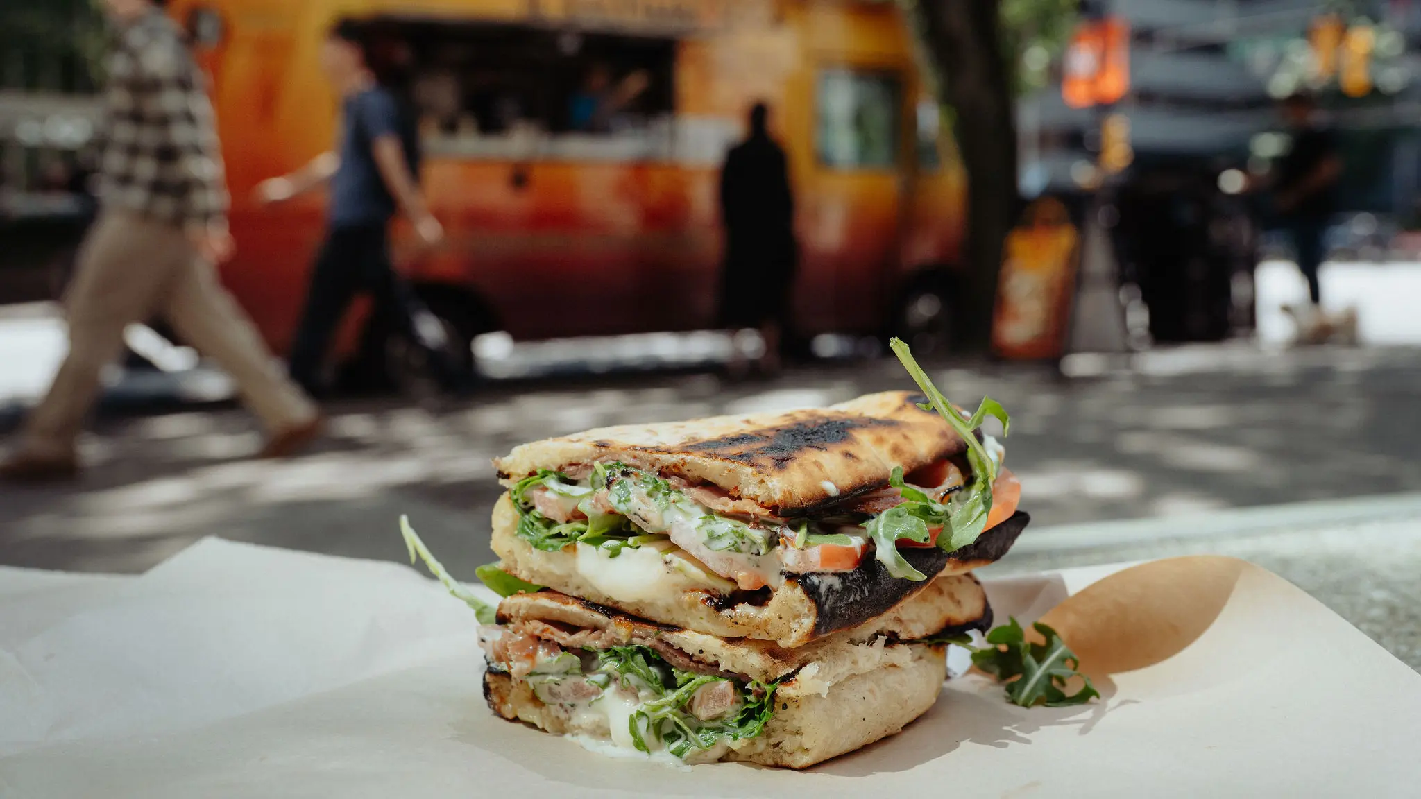 Close-up of a gourmet sandwich with arugula and savory toppings, cut in half to reveal its layers, with the colorful Il Saltimbocca food truck parked in downtown Vancouver in the background.