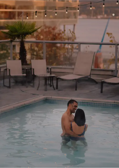 A couple embraces in a rooftop pool surrounded by lounge chairs and string lights, overlooking the cityscape and water at sunset—discover romantic Vancouver attractions with exclusive discounts for an unforgettable evening.