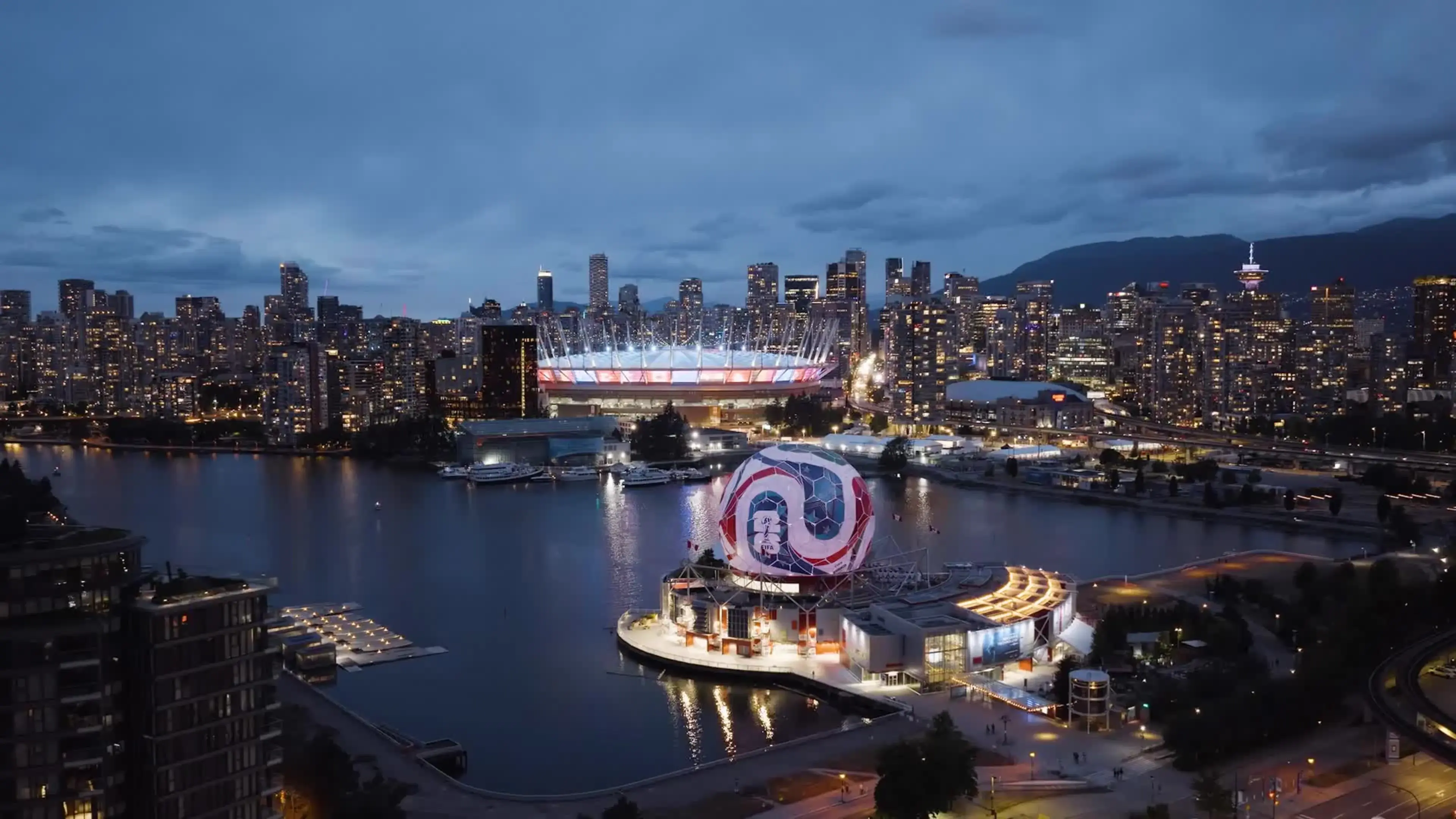 Night view of a city skyline with illuminated buildings, a stadium, and a waterfront area with a large spherical structure.