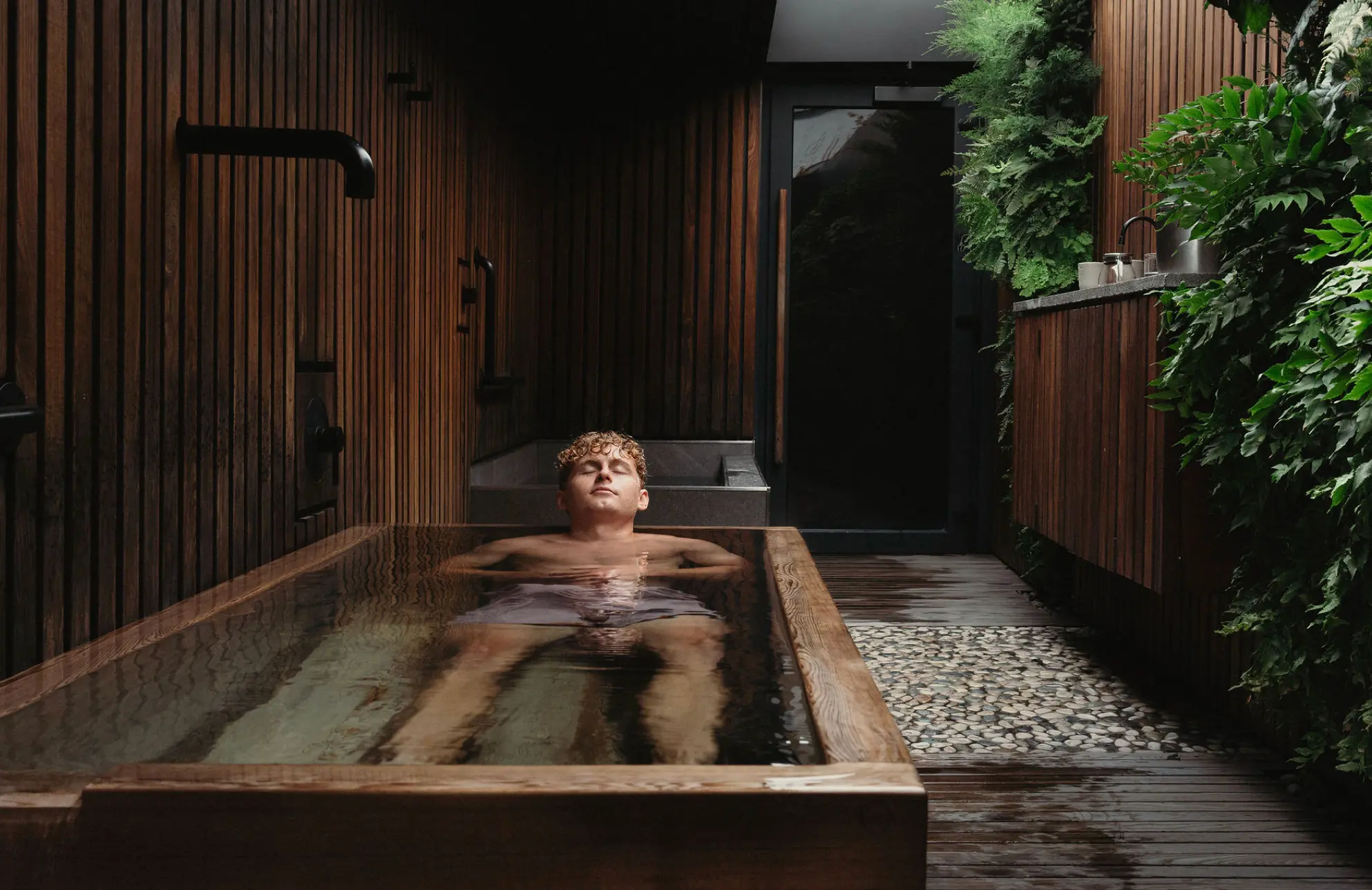 Person relaxing in a wooden bathtub in a spa-like room with dark wood walls and green plants.