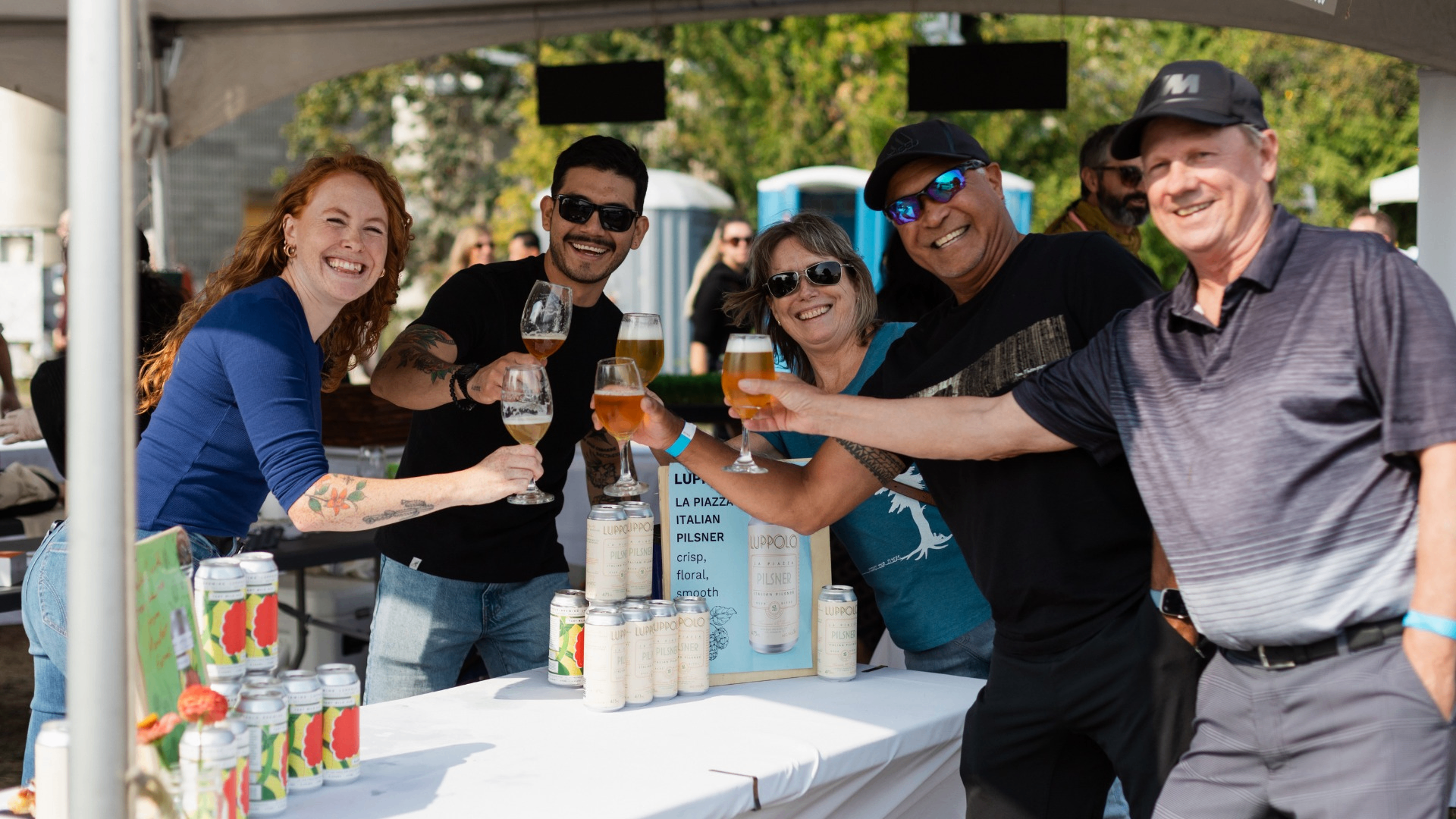 Five people smiling and toasting with glasses of beer at an outdoor event booth with cans and a sign on the table.