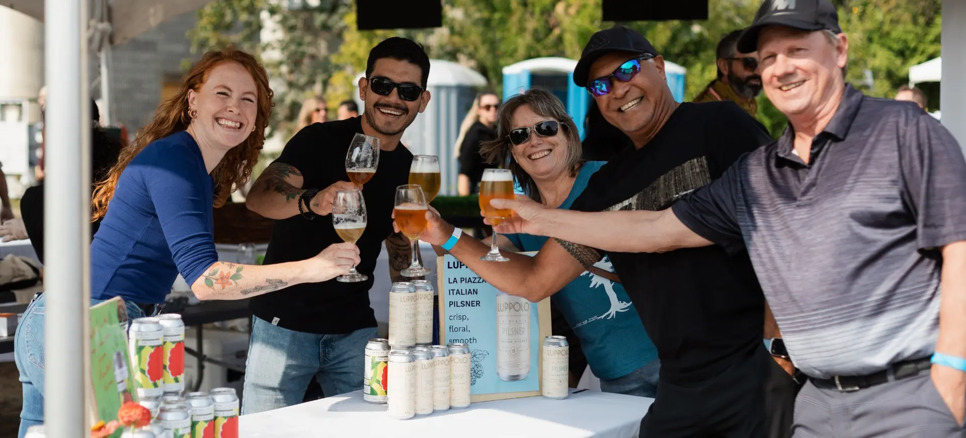 Five people smiling and toasting with glasses of beer at an outdoor event booth with cans and a sign on the table.