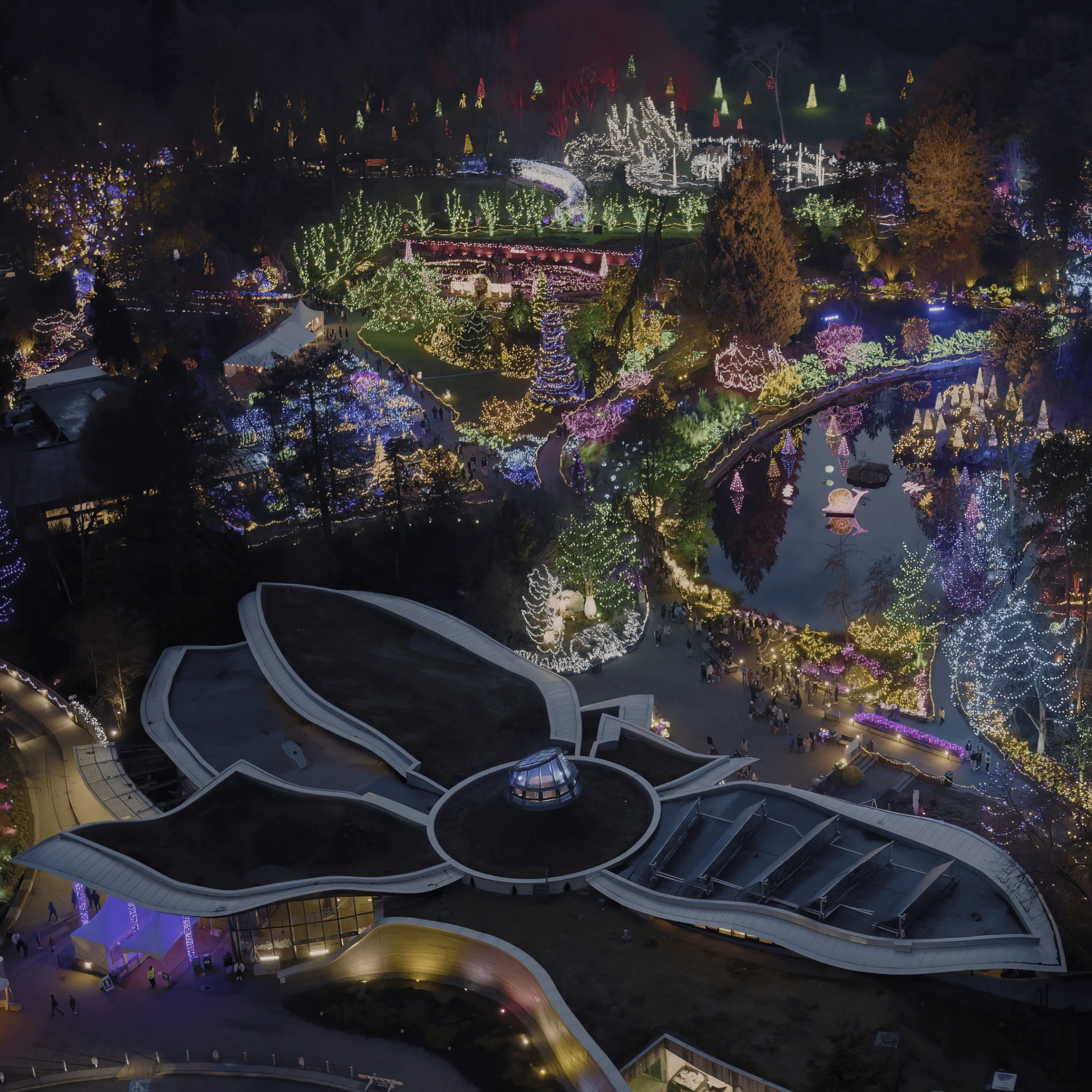 An aerial view of a garden at night, illuminated with colorful holiday lights outlining trees, pathways, and structures, with crowds of people enjoying the festive display—a must-see highlight in any Vancouver travel guide.