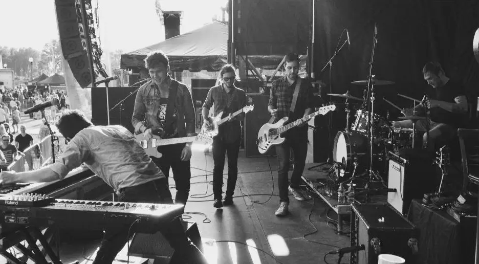 Black and white photo of a five-member band performing on an outdoor stage with guitars, keyboard, and drums.