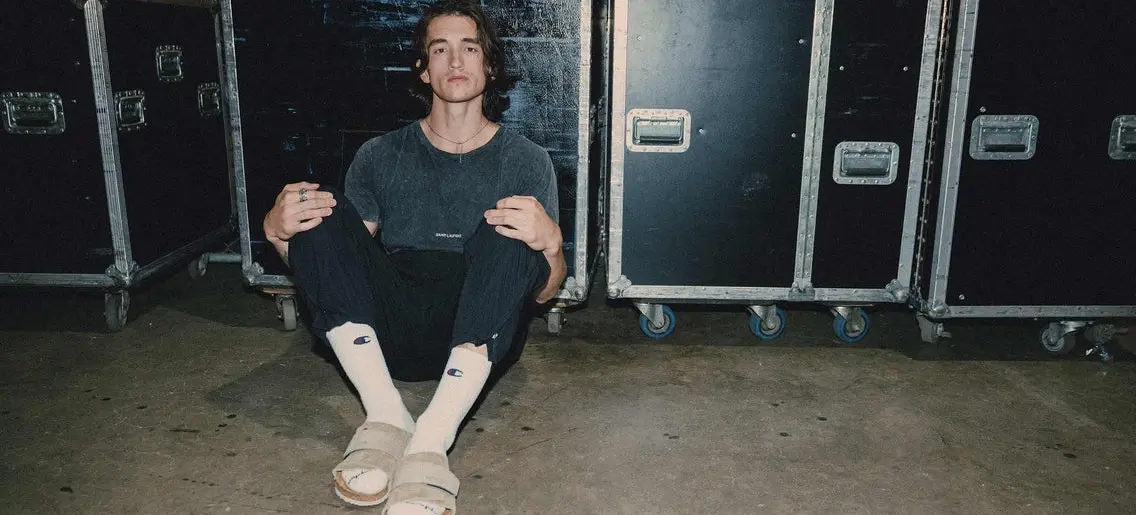 A person sitting on the floor with knees up, wearing a dark shirt, black pants, white socks, and beige sandals, against a backdrop of black equipment cases.