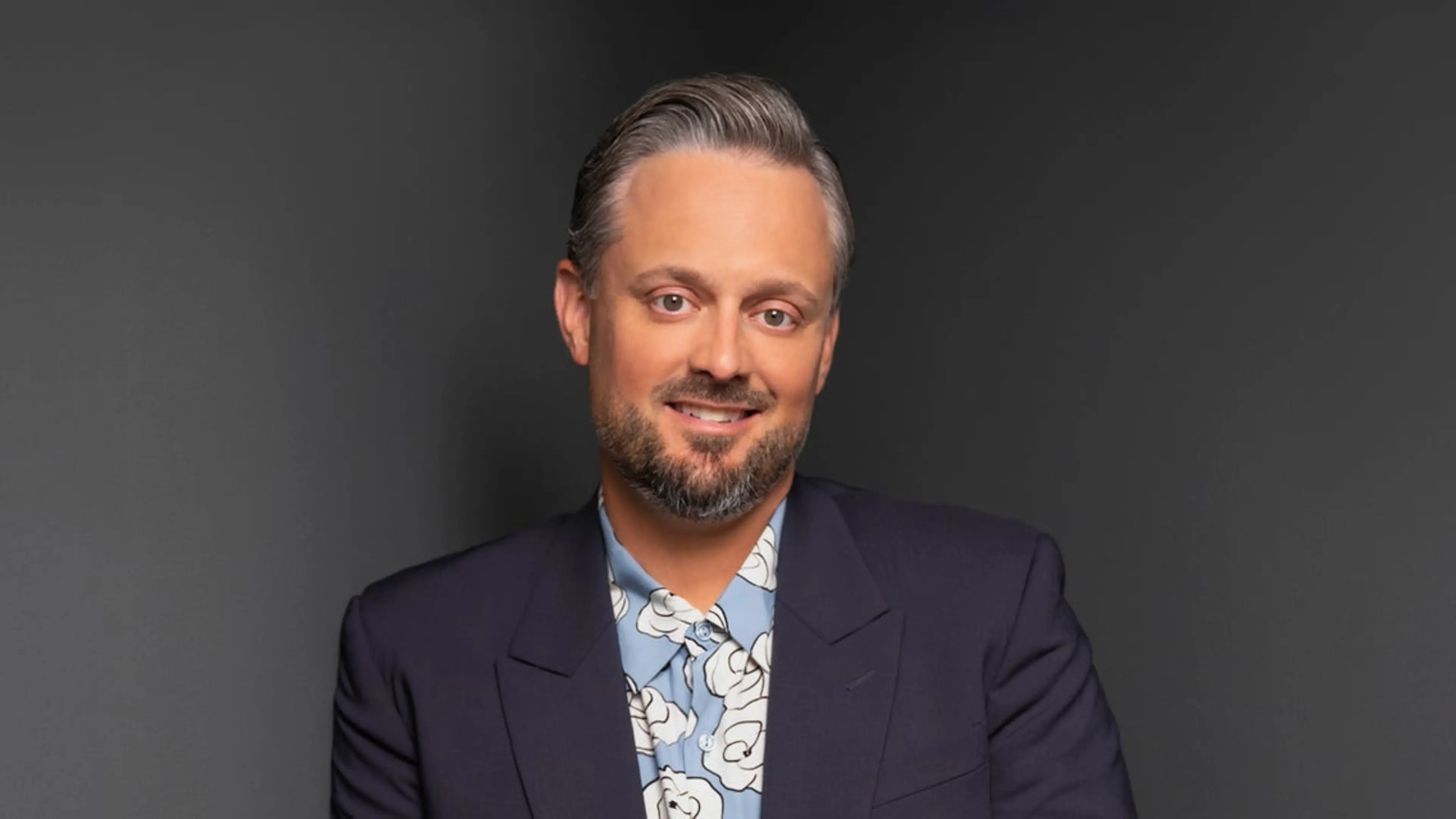 Person with short gray hair and beard wearing a dark blazer and blue floral shirt, smiling against a dark background.