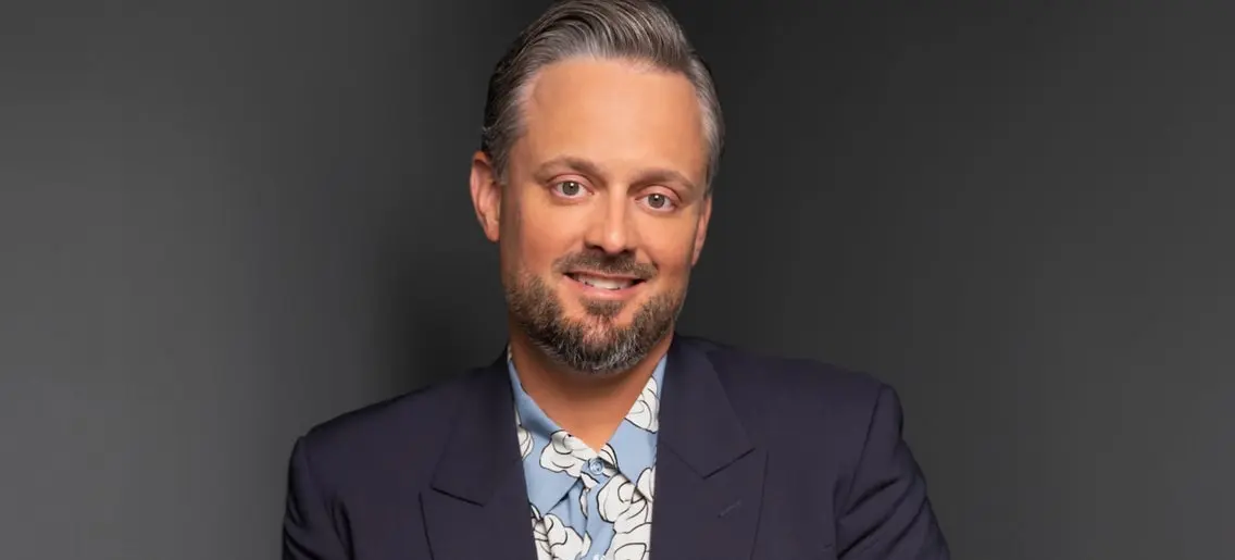 Person with short gray hair and beard wearing a dark blazer and blue floral shirt, smiling against a dark background.