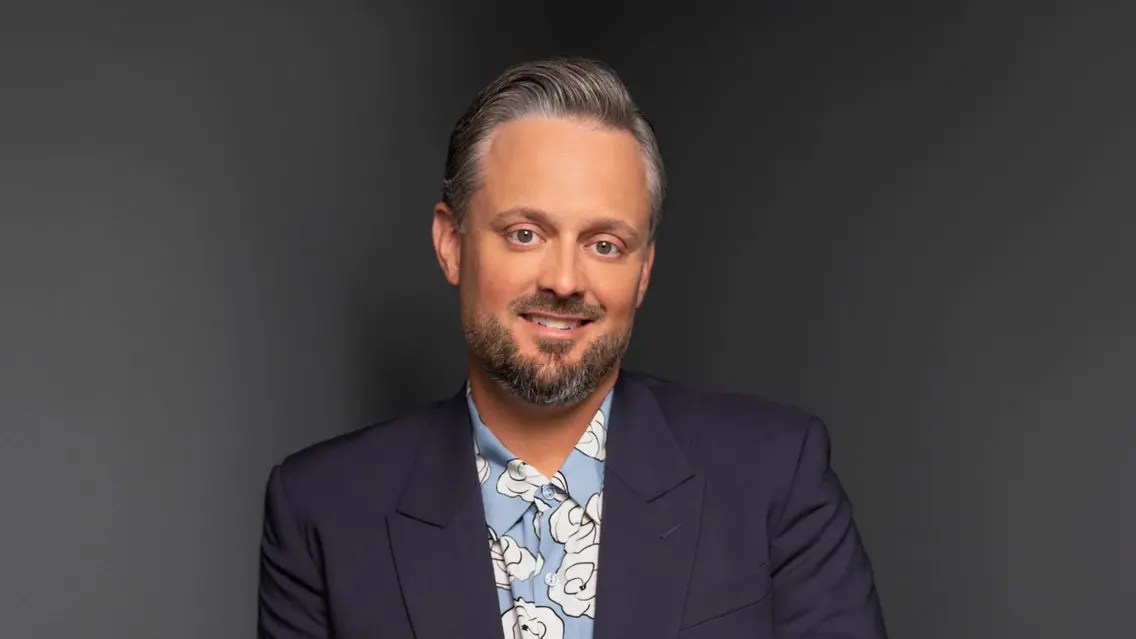 Person with short gray hair and beard wearing a dark blazer and blue floral shirt, smiling against a dark background.