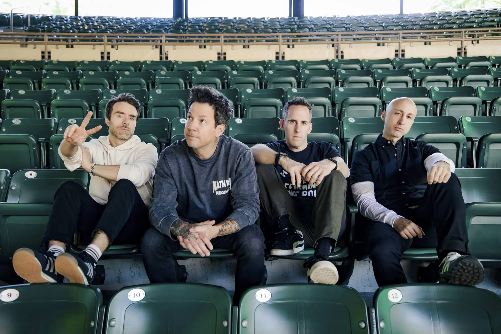 Four men sitting in green stadium seats, one making a peace sign, others looking forward or to the side.