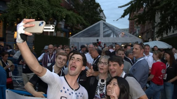 A group of five people smiling and posing for a selfie at an outdoor event with a crowd and a tent in the background.