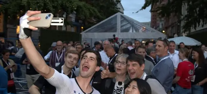 A group of five people smiling and posing for a selfie at an outdoor event with a crowd and a tent in the background.
