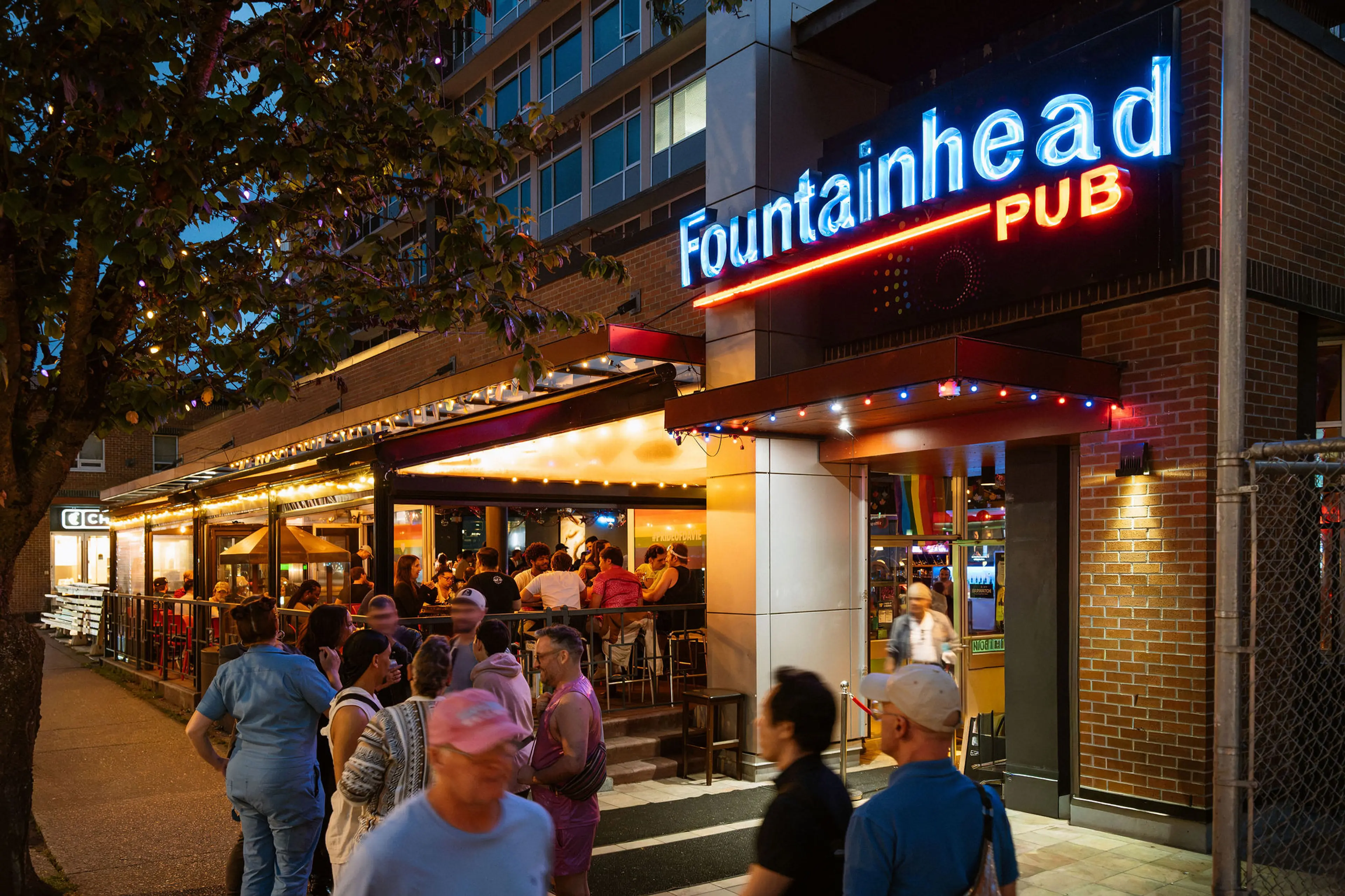 Crowd of people outside Fountainhead Pub at night with neon sign and outdoor seating area.