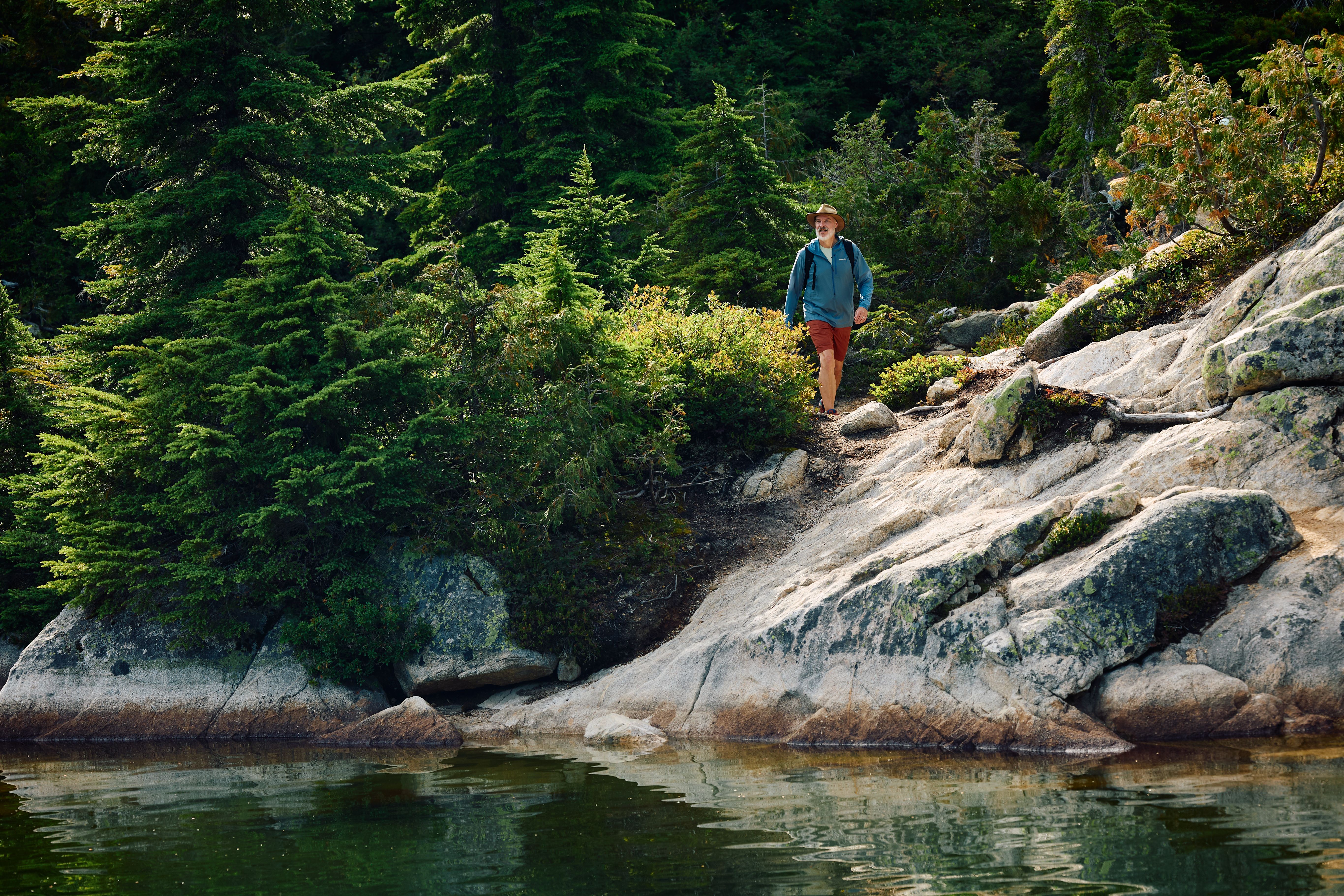 Man wearing a hat, blue jacket, and red shorts hiking on a rocky trail near a forested lake.