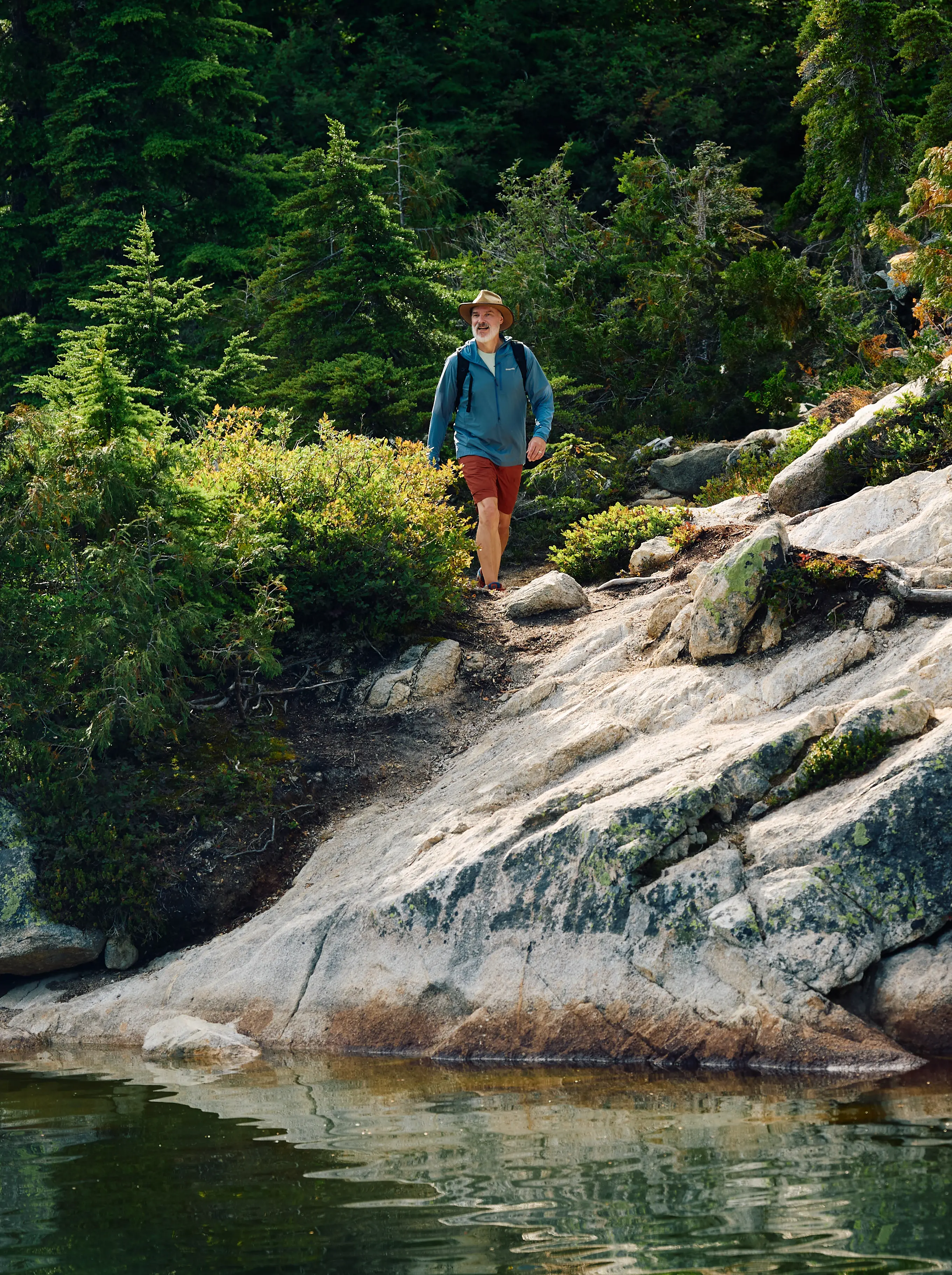 Man wearing a hat, blue jacket, and red shorts hiking on a rocky trail near a forested lake.