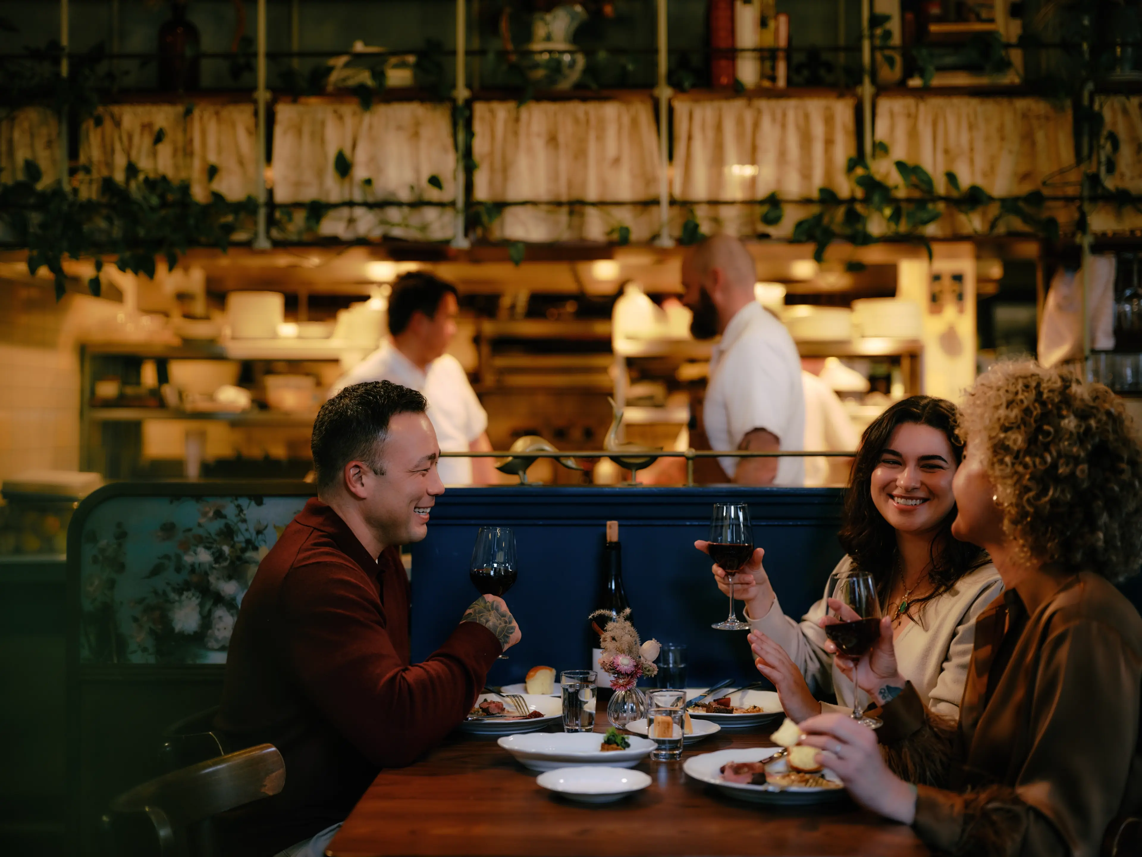 Three people sitting at a restaurant table, smiling and holding wine glasses, with chefs working in the kitchen behind them at the St. Lawrence restaurant in Vancouver.