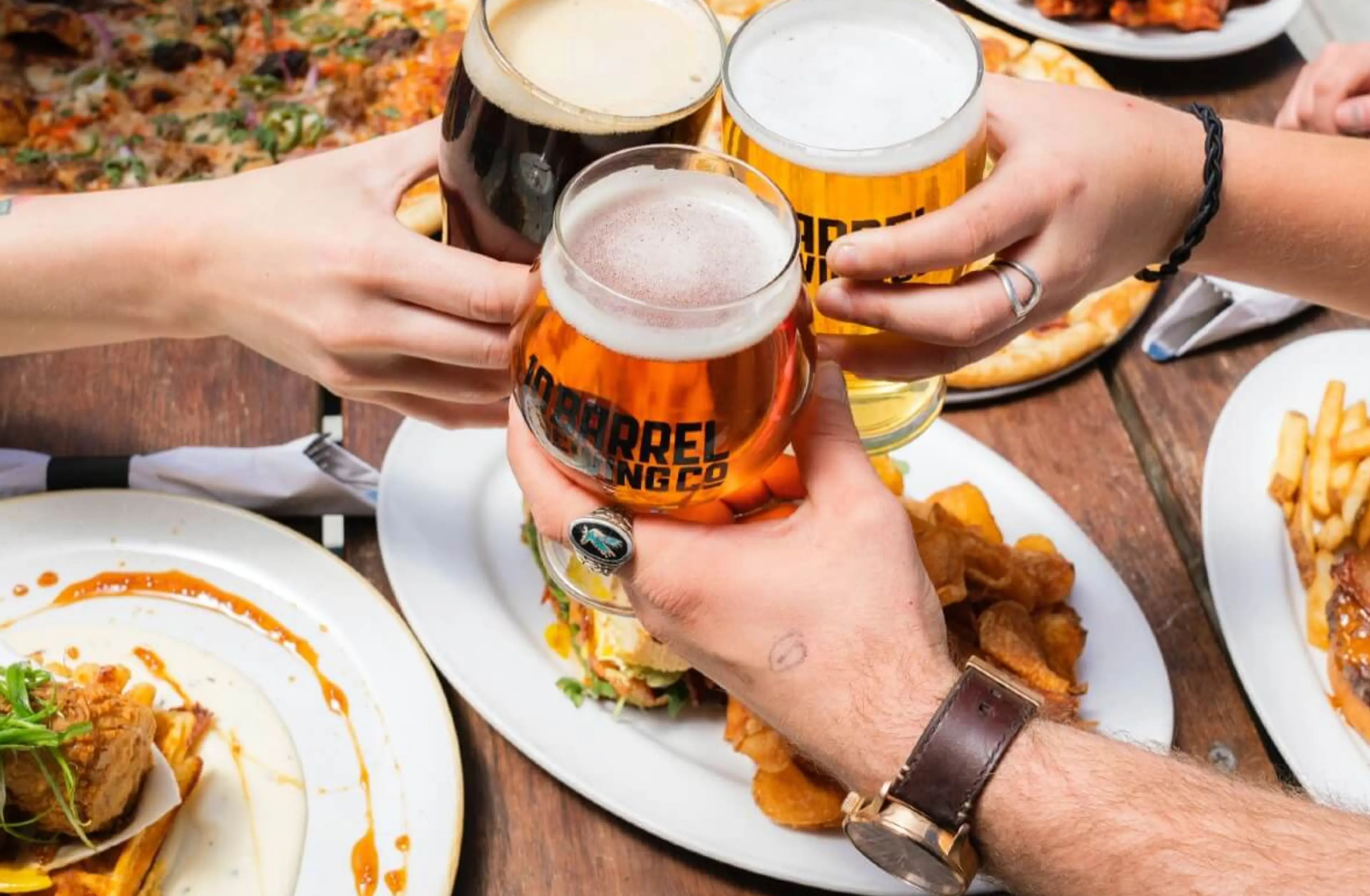 Three people clinking glasses of beer over plates of food including pizza, fries, and a sandwich on a wooden table.