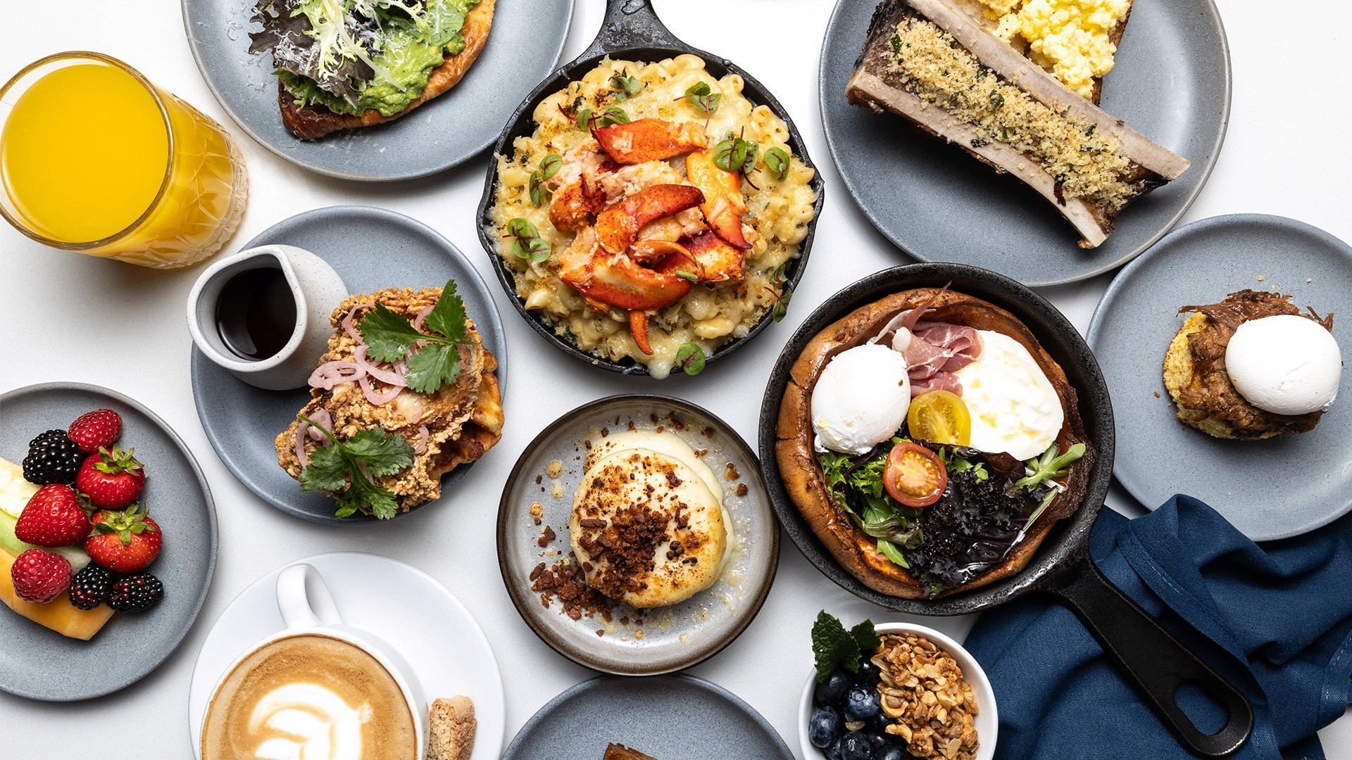 Assorted breakfast dishes including eggs, toast, fruit, coffee, orange juice, and a skillet with salad and poached eggs on a white table.
