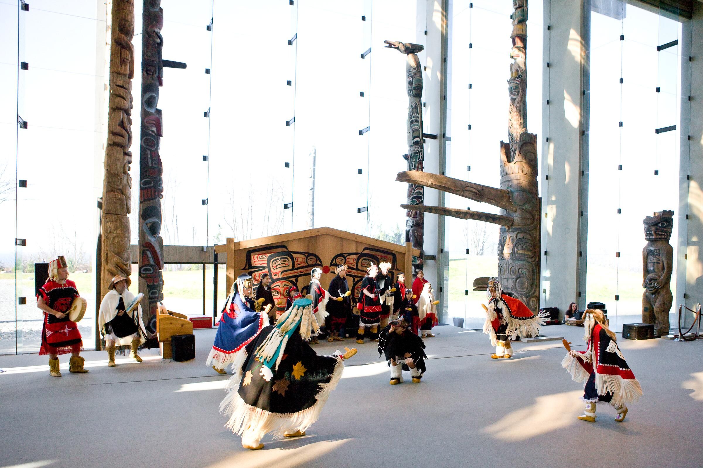 Group of people in traditional Indigenous clothing performing a dance inside a building with large glass windows and tall totem poles.