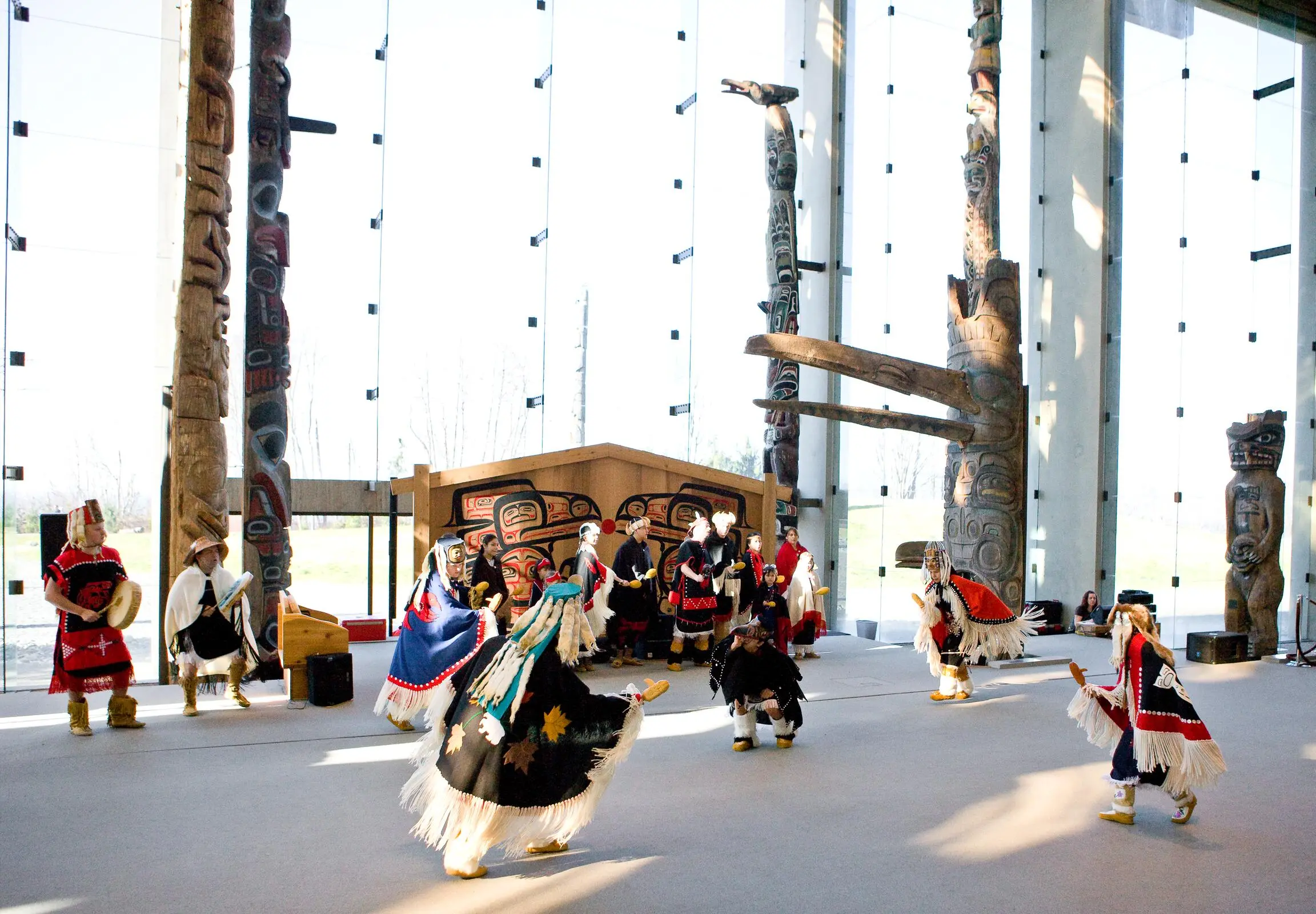 Group of people in traditional Indigenous clothing performing a dance inside a building with large glass windows and tall totem poles.