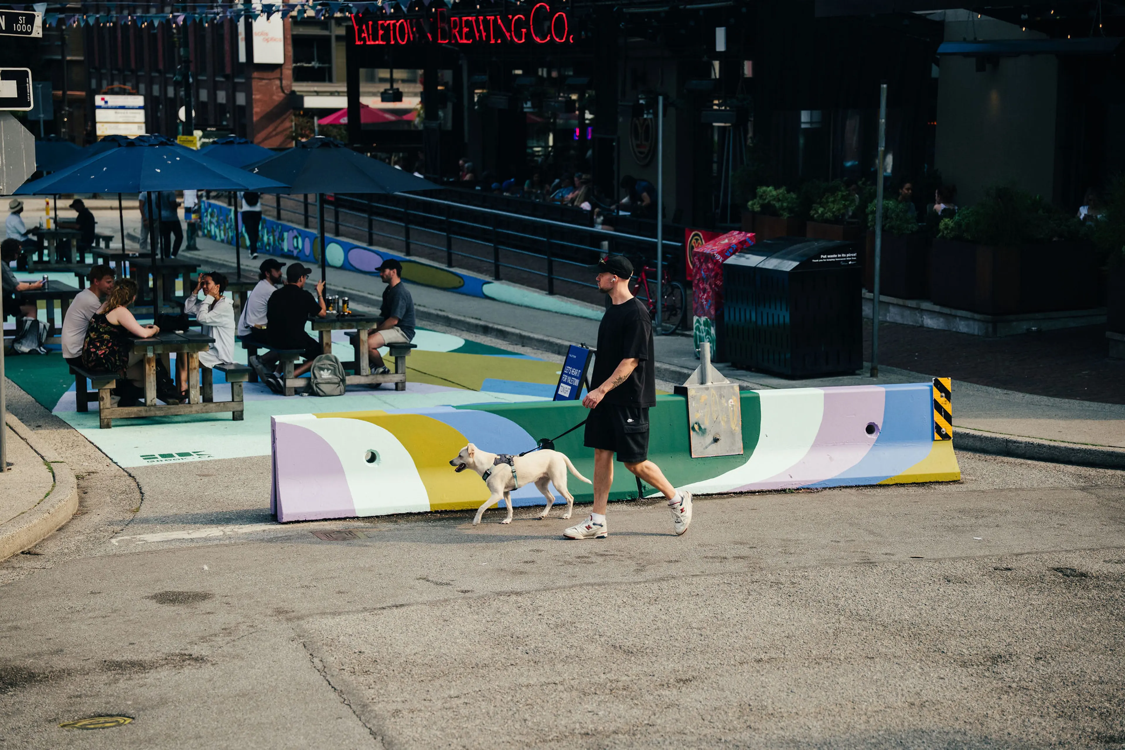 Man in black outfit walking a white dog on a leash near colorful street barriers with people sitting at picnic tables under umbrellas.