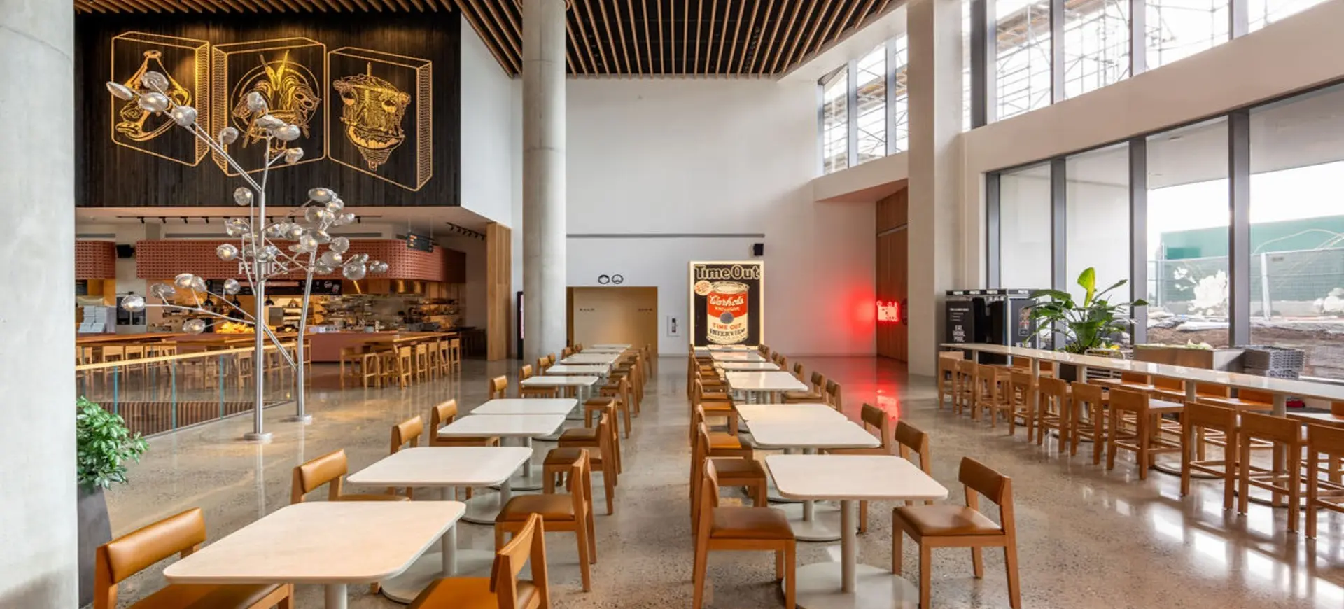 Empty modern cafeteria with rows of tables and chairs, large windows on the right, and decorative light fixtures.