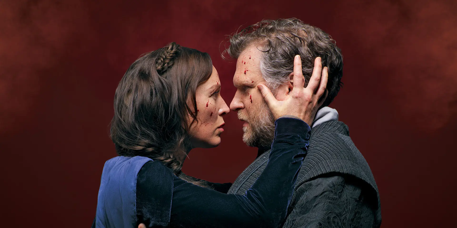 Two people with blood on their faces looking intensely at each other, one holding the other's face against a dark red background.