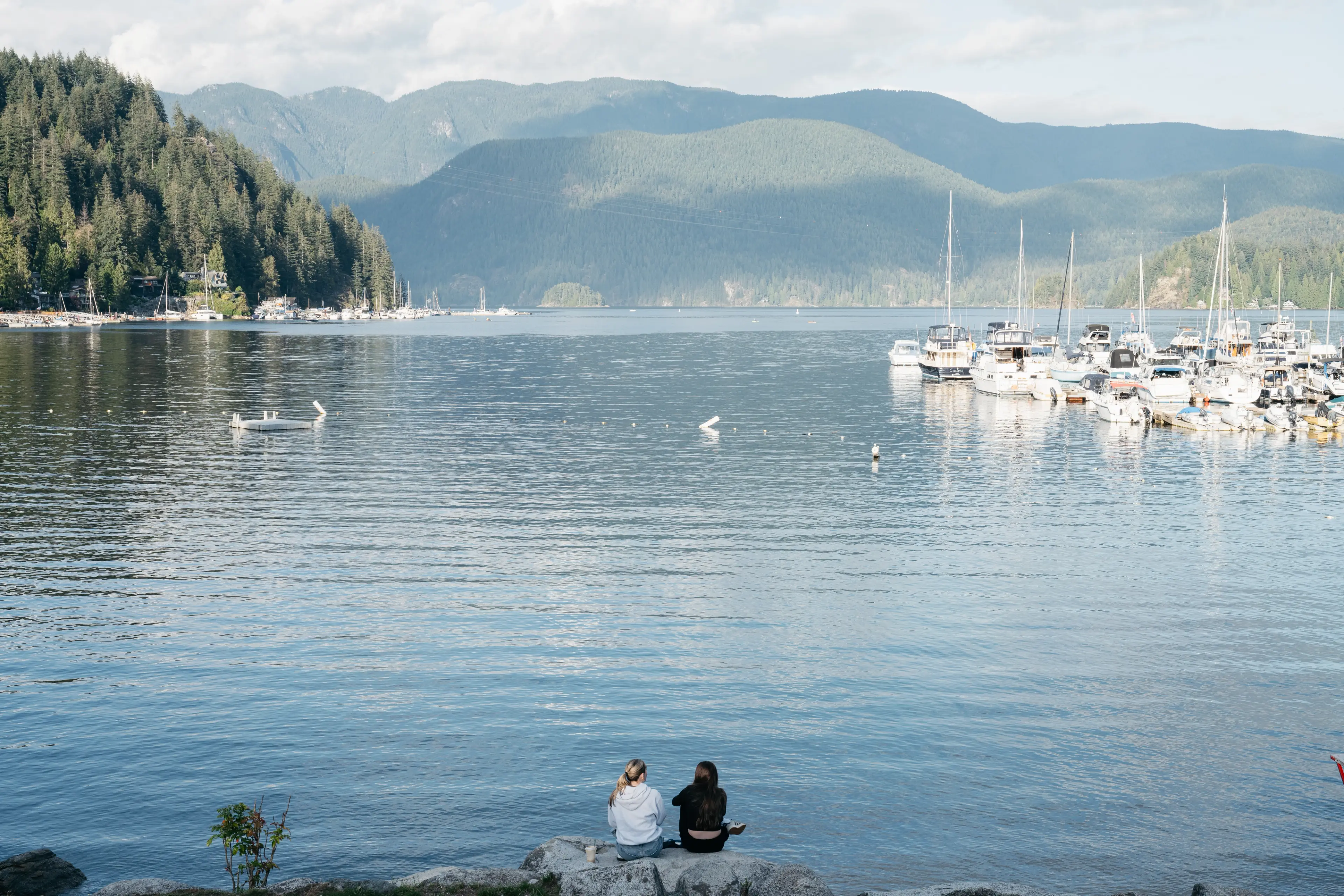 Two people sitting on rocks by a calm lake with boats docked on the right and forested mountains in the background.