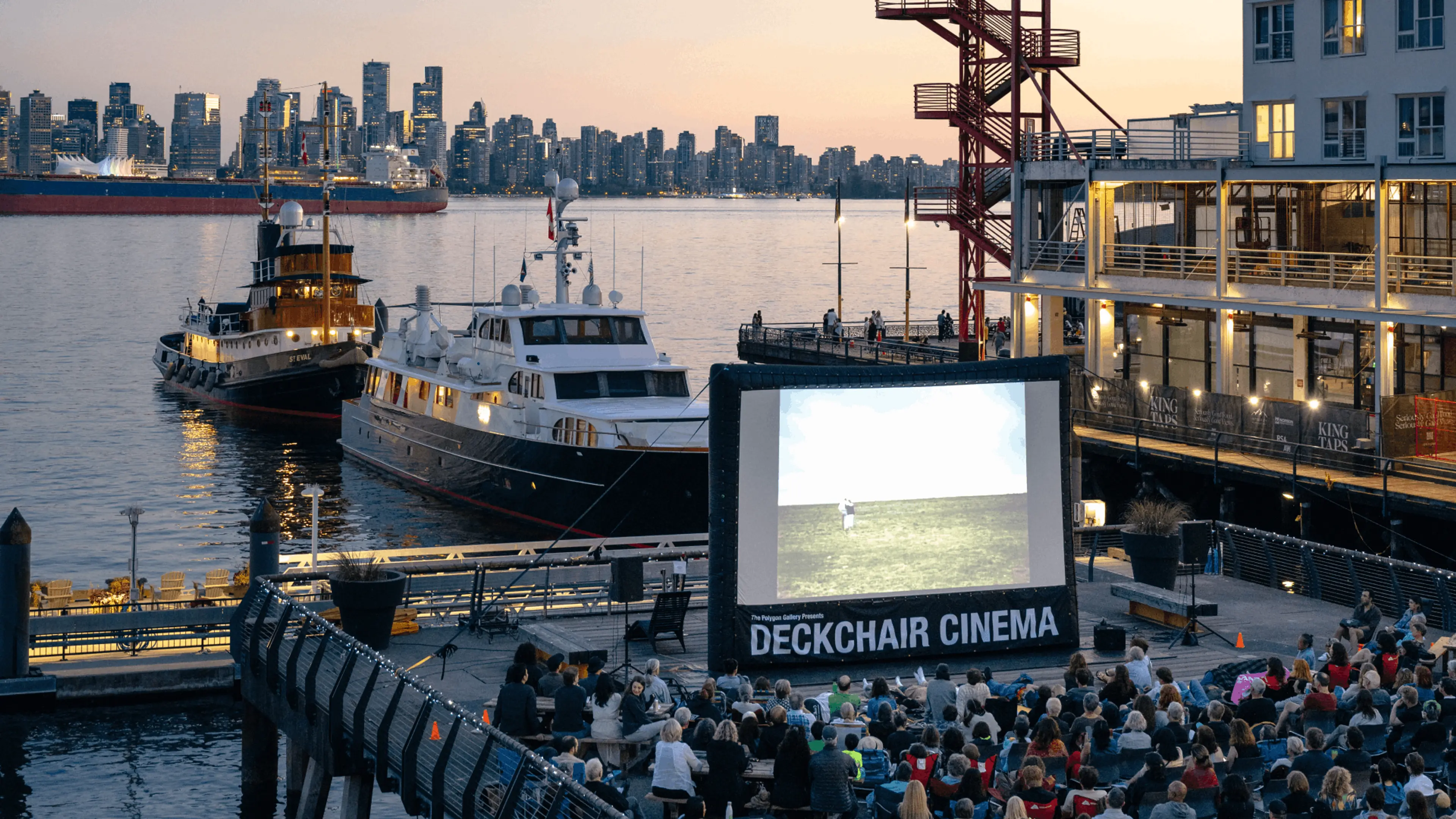 An outdoor "Deckchair Cinema" event at The Polygon Gallery in North Vancouver, featuring a large inflatable movie screen on the waterfront with the downtown Vancouver skyline across the harbor.