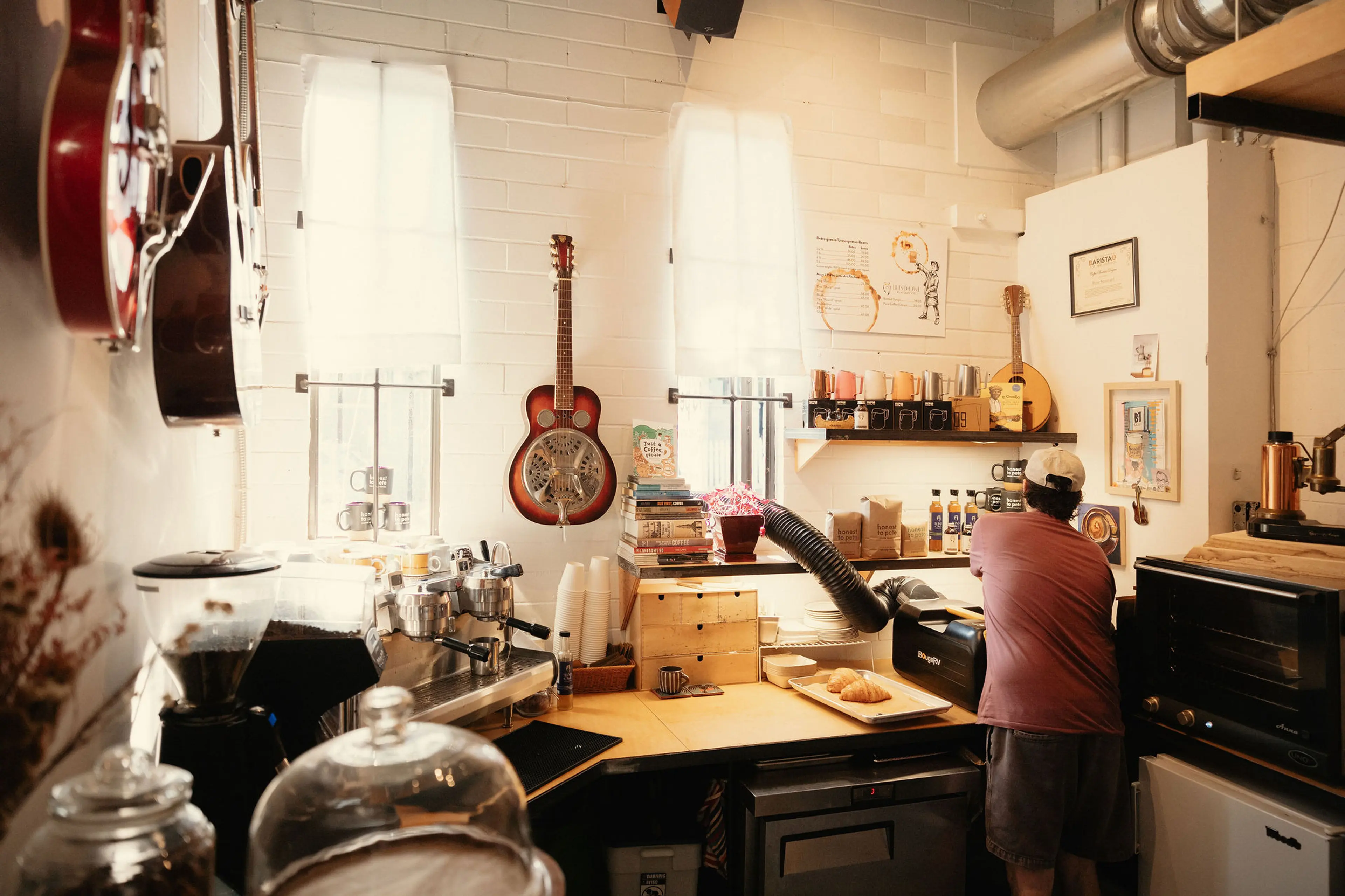 Person working behind a counter in a cozy cafe with guitars on the wall, coffee equipment, and pastries on the counter.