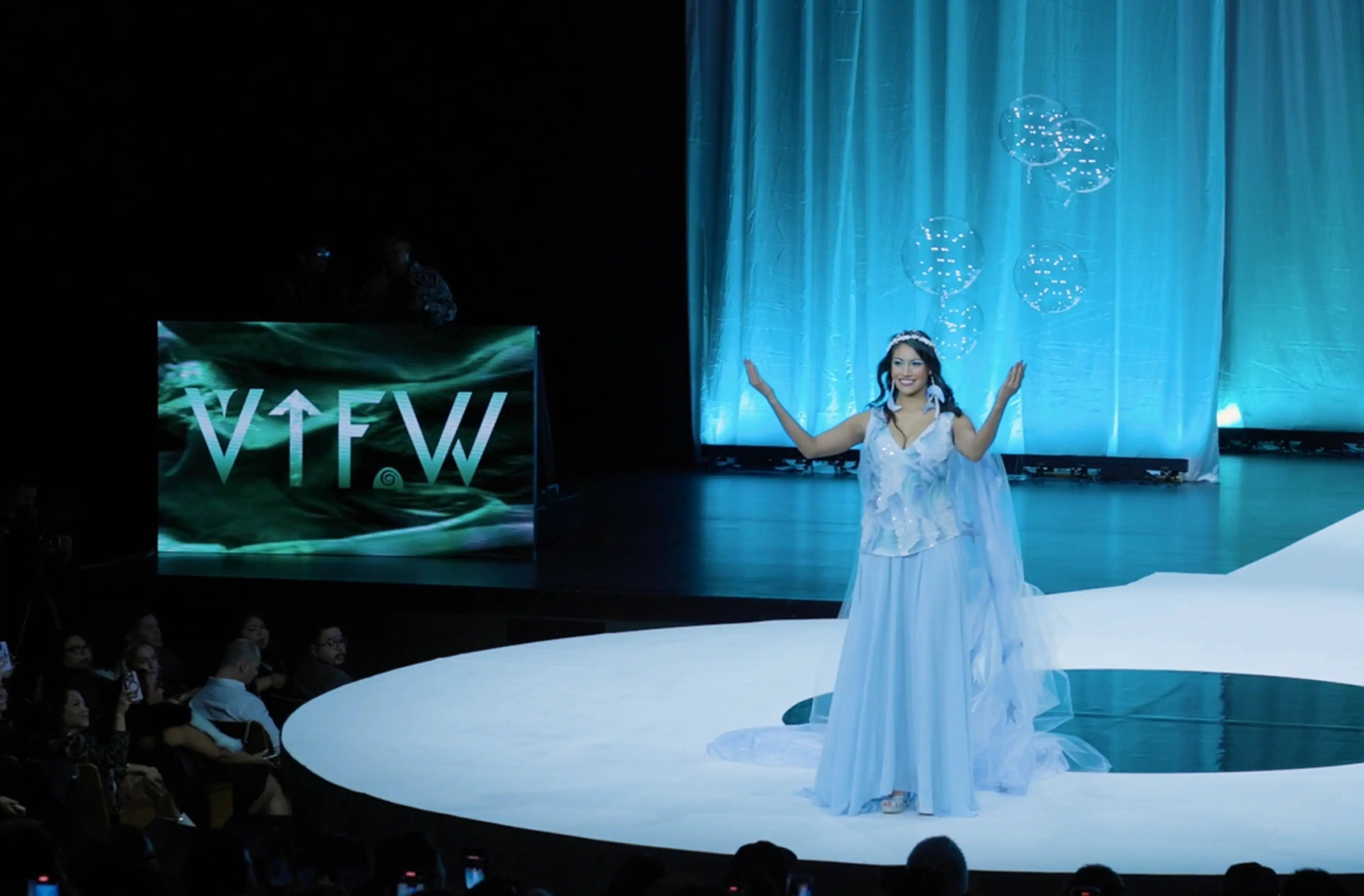 Person in a light blue gown and tiara on a runway stage with floating transparent balloons and a blue curtain backdrop.