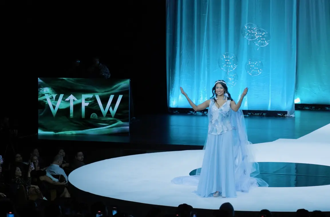 Person in a light blue gown and tiara on a runway stage with floating transparent balloons and a blue curtain backdrop.
