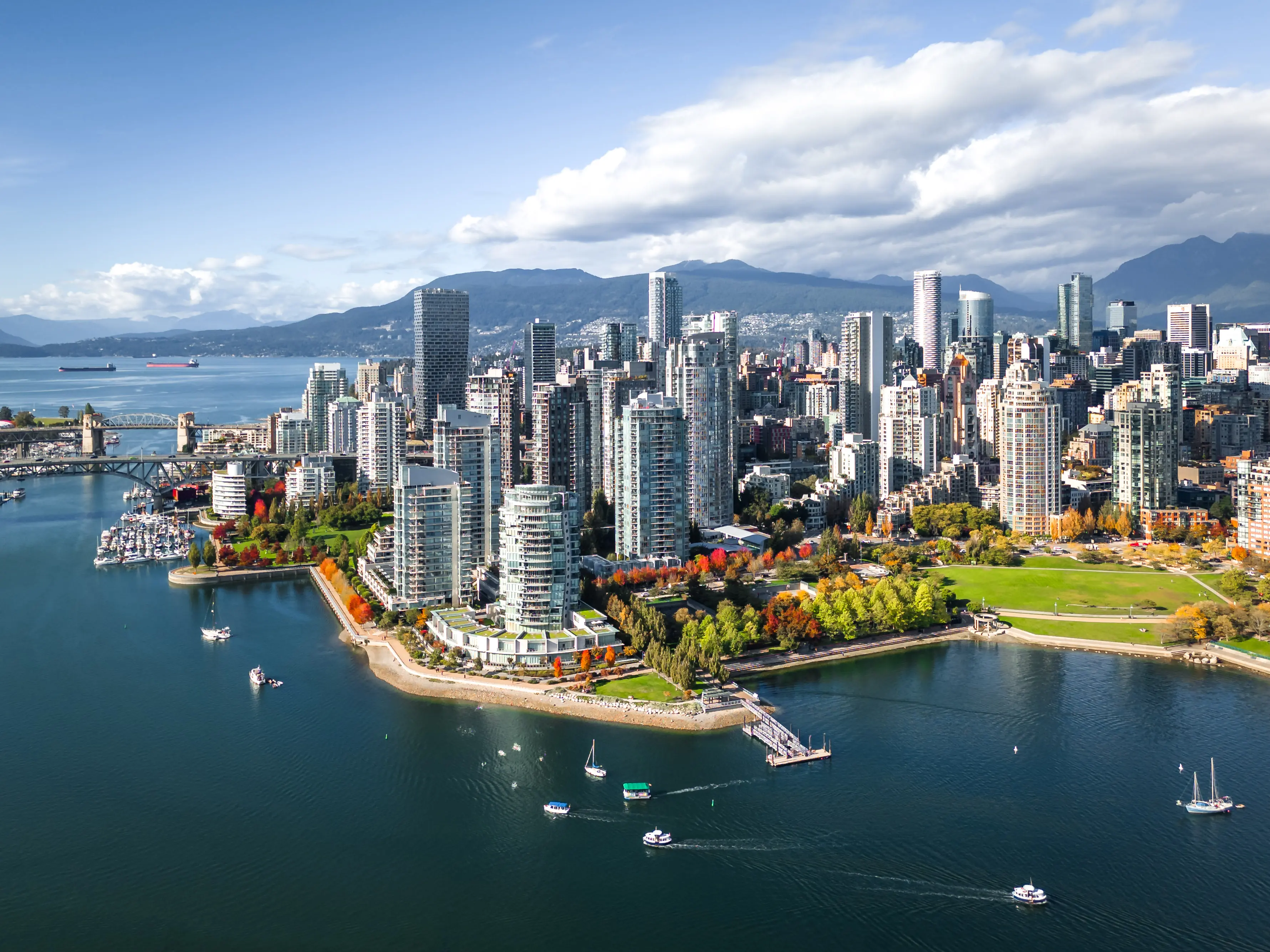 Aerial view of a coastal city with high-rise buildings, green parks, boats on the water, and mountains in the background.