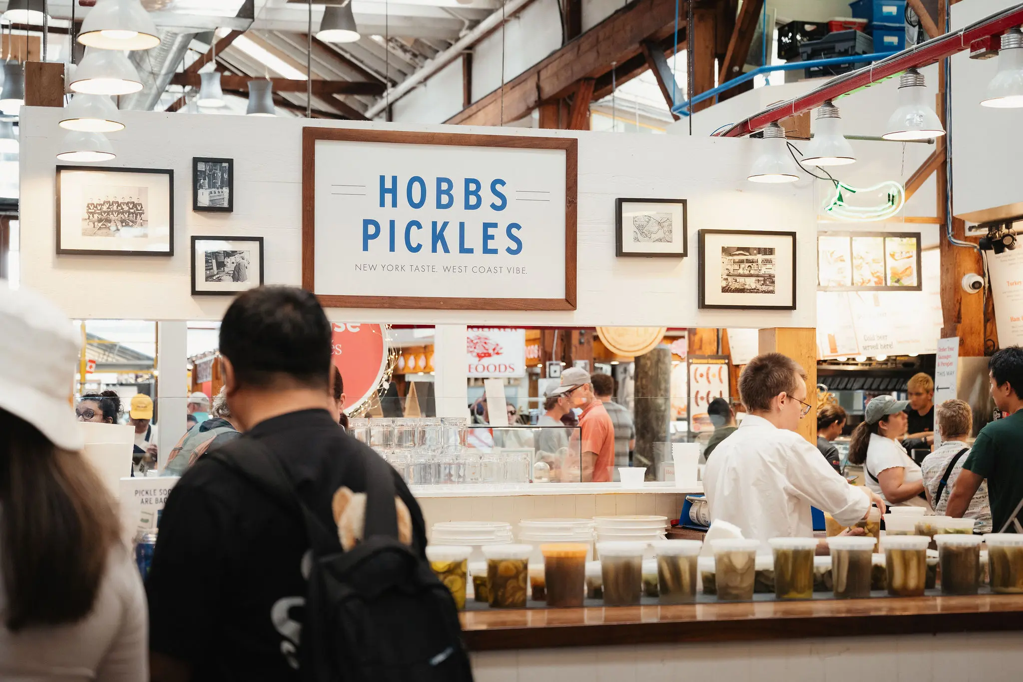 People at a market stall named Hobbs Pickles with jars of pickles lined up on the counter.