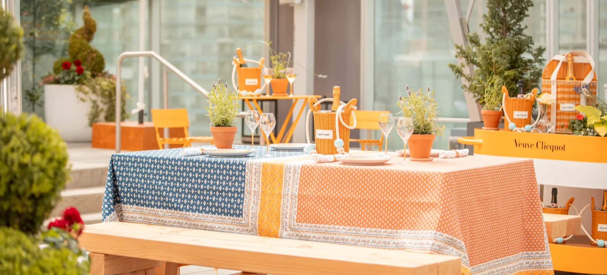 Outdoor dining table with blue and orange patterned tablecloth, potted plants, wine glasses, and wooden bench under umbrellas.
