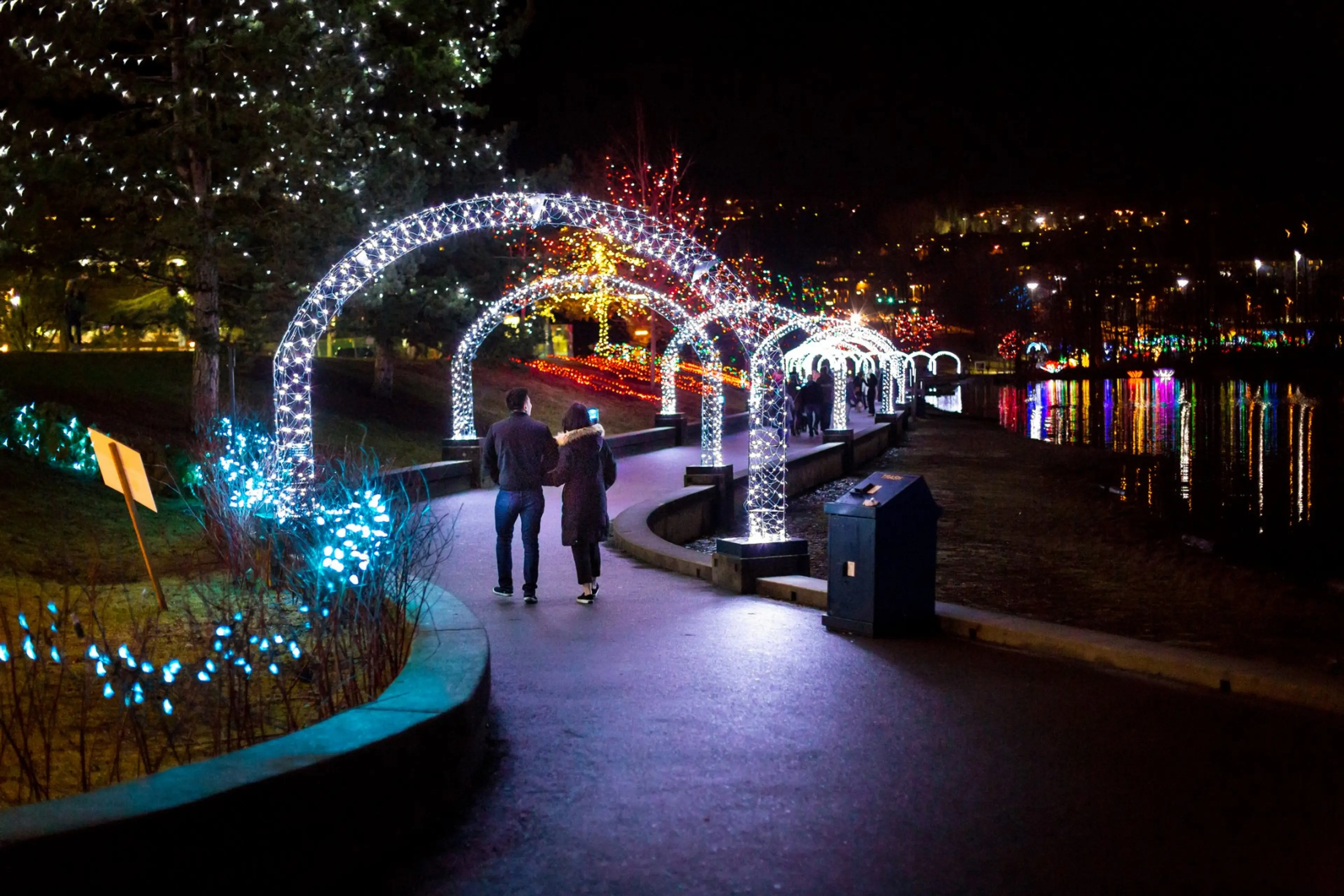 A couple walks along a path beside Lafarge Lake in Coquitlam. They are walking beneath a lighted arch and there are holiday lights set up as part of the Lights at Lafarge event.