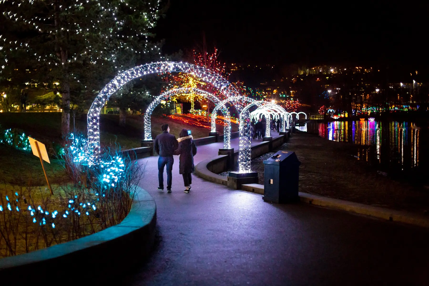 A couple walks along a path beside Lafarge Lake in Coquitlam. They are walking beneath a lighted arch and there are holiday lights set up as part of the Lights at Lafarge event.