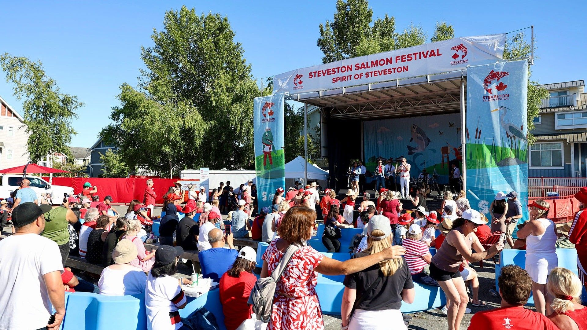 Crowd of people seated and standing in front of a stage at Steveston Salmon Festival on a sunny day.