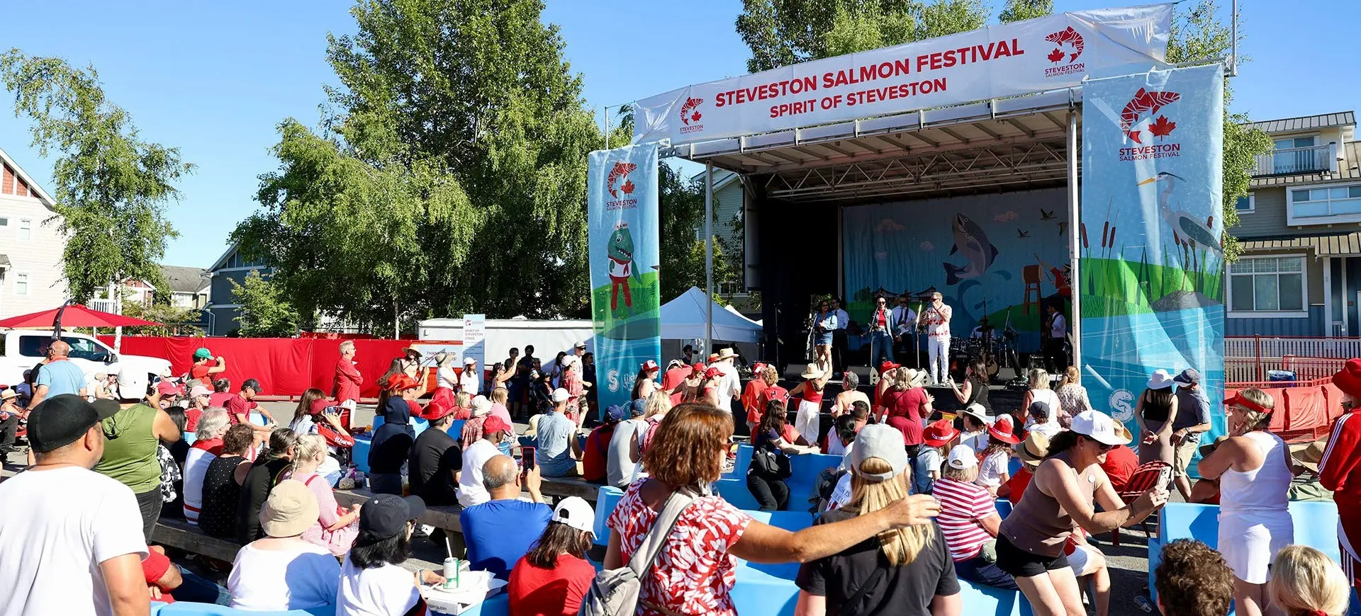 Crowd of people seated and standing in front of a stage at Steveston Salmon Festival on a sunny day.