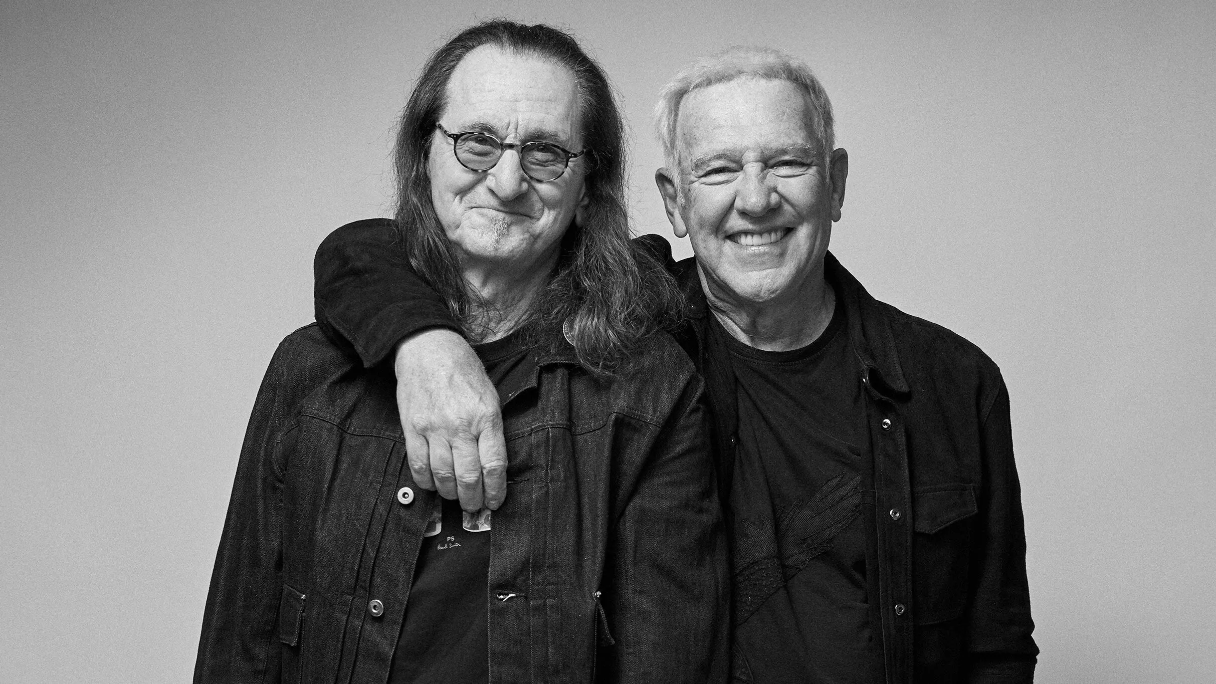 Two older people smiling, one with long hair and glasses, the other with short hair, arm around the other's shoulder.