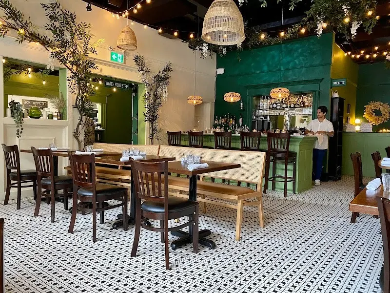 Empty restaurant interior with wooden chairs, tables set with glasses and napkins, green walls, hanging lights, and a person behind the bar.