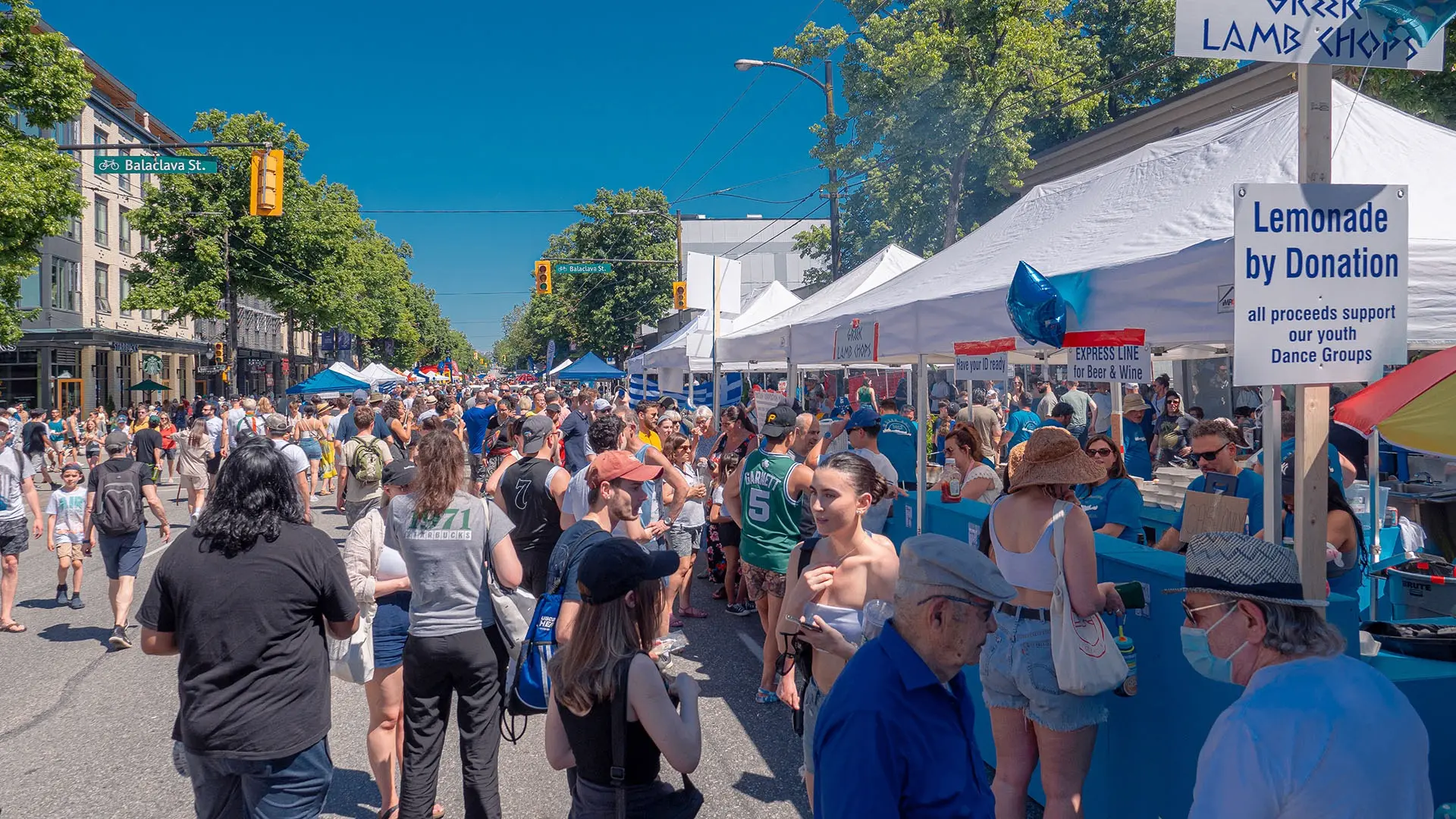 Crowded street fair with people walking and standing near white tents, one tent has a sign for lemonade by donation.