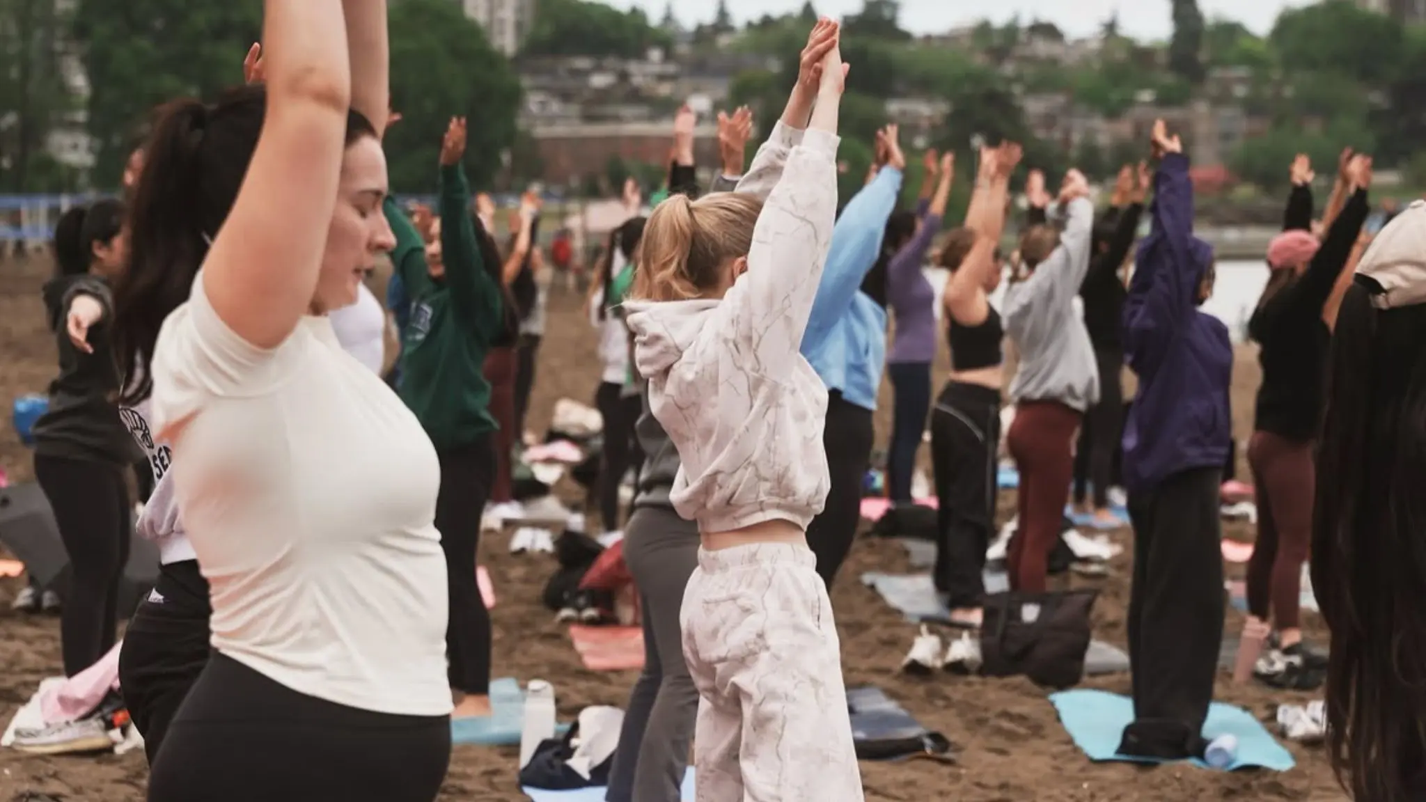 A group of people standing on mats outdoors with arms raised, participating in a group exercise or yoga session.