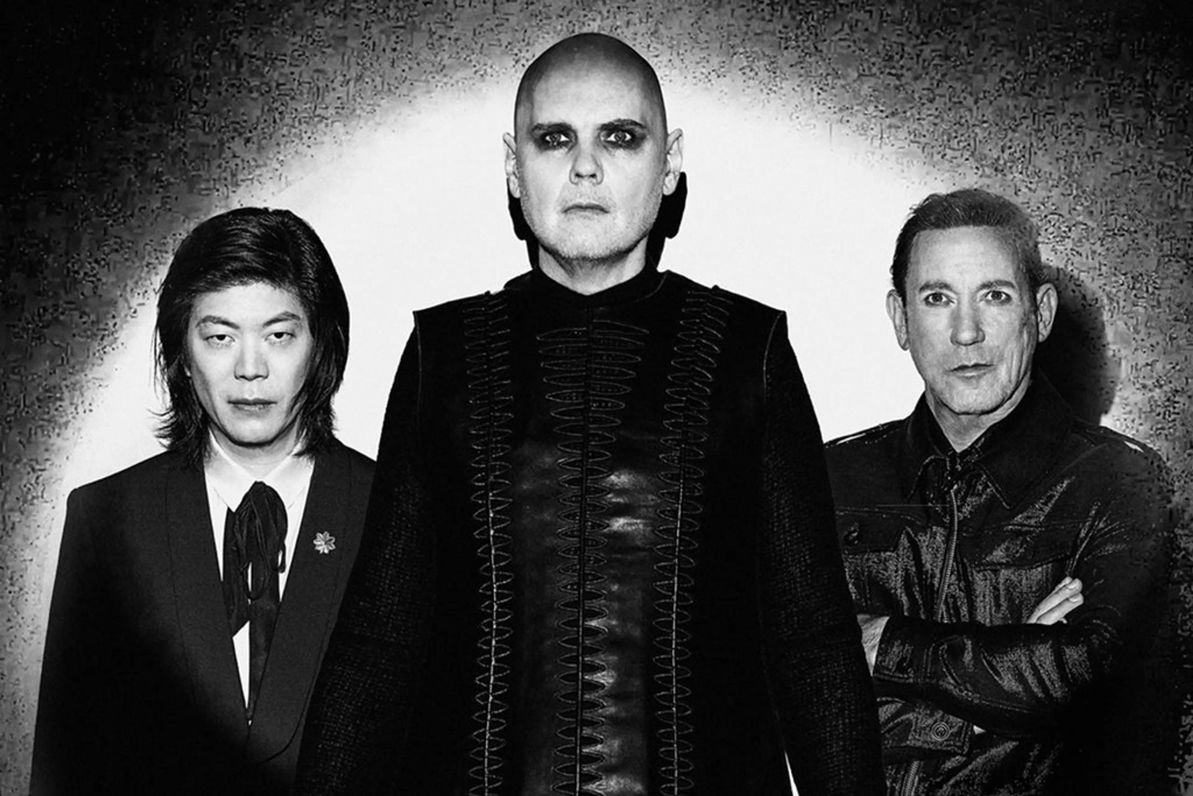 Three people standing against a wall with spotlight; center person is bald with dark eye makeup and black outfit, others wear dark jackets.