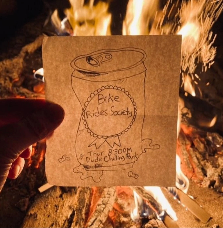 Hand holding a paper with a drawing of a soda can and text reading 'Bike Rides Society, Thur 8:30 PM, Dude Chilling Park' in front of a campfire.