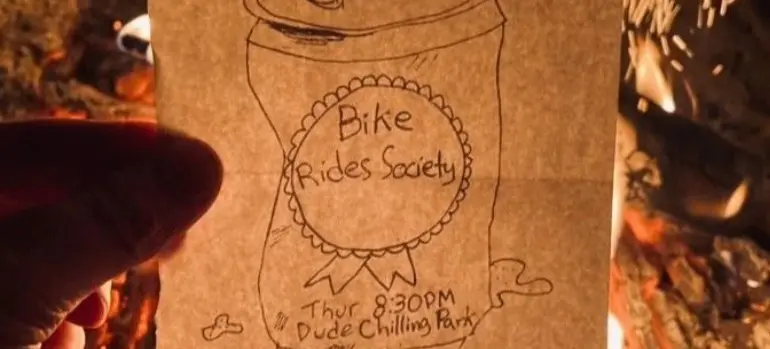 Hand holding a paper with a drawing of a soda can and text reading 'Bike Rides Society, Thur 8:30 PM, Dude Chilling Park' in front of a campfire.