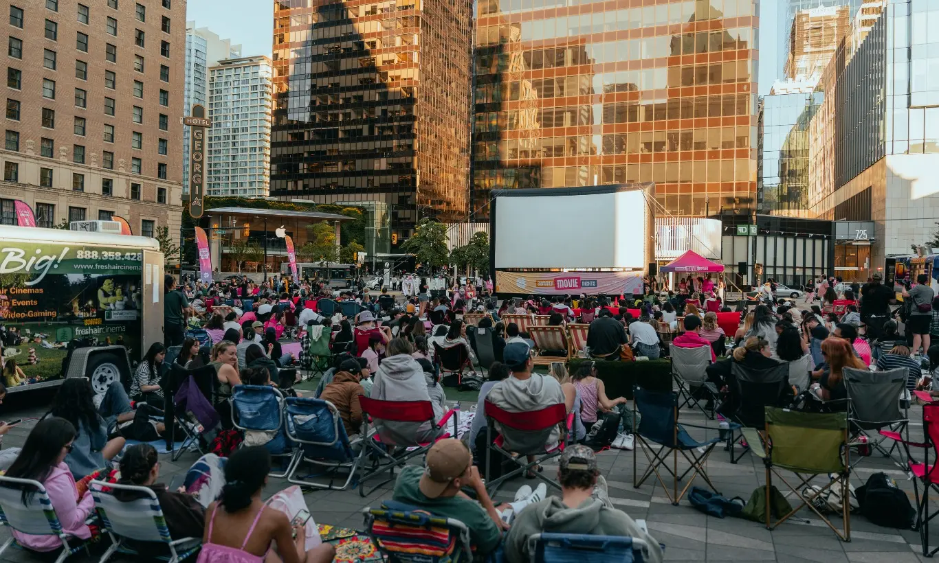 Large crowd seated on folding chairs and blankets in an urban plaza watching an outdoor movie on a large screen with tall buildings in the background.