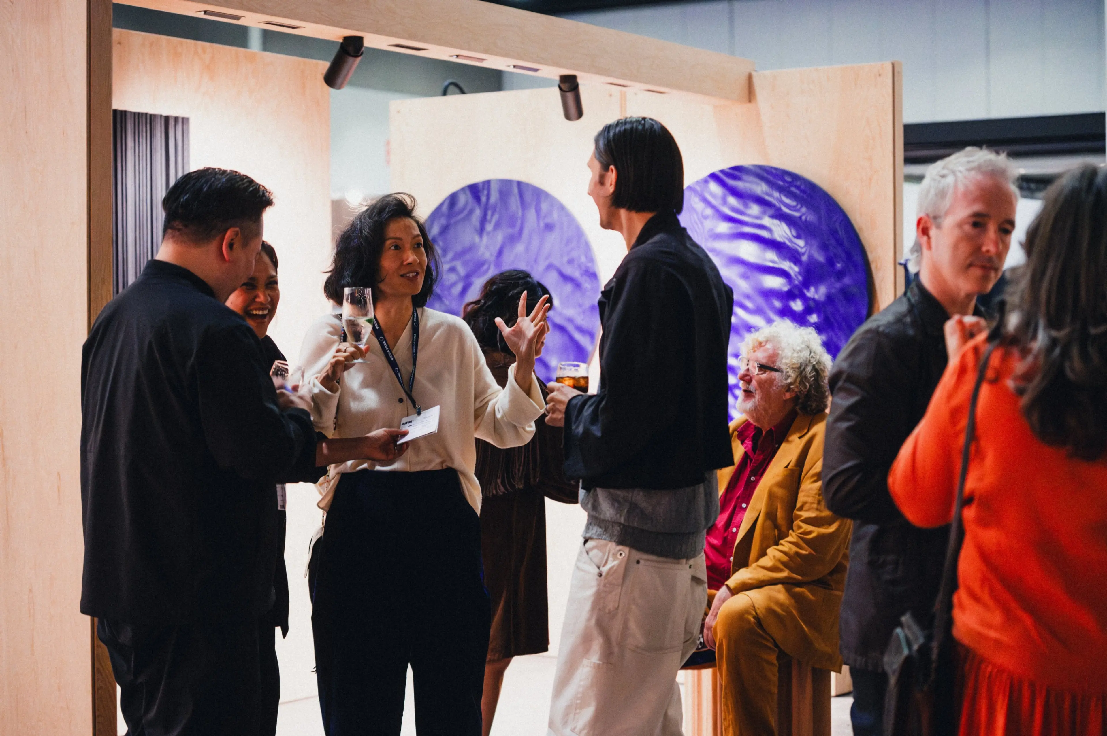 A group of people socializing indoors, some holding drinks, with two large blue circular artworks on the wall behind them.