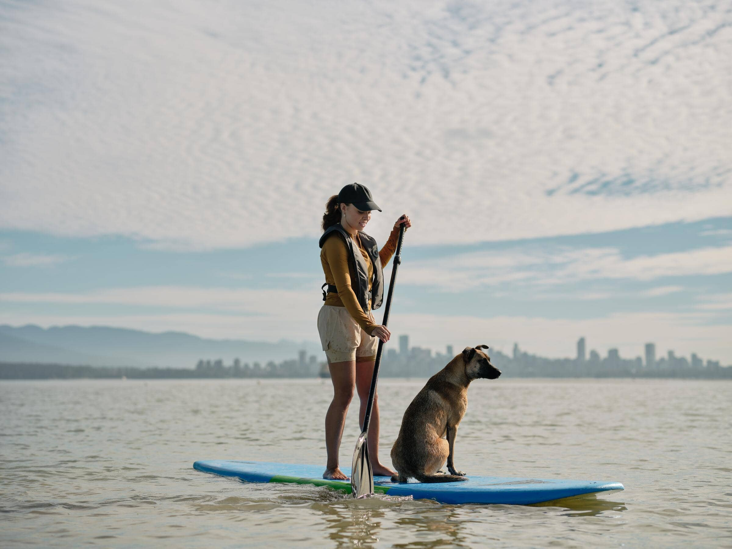 Woman in a cap and life jacket paddleboarding on calm water with a dog sitting on the board, city skyline in the background.